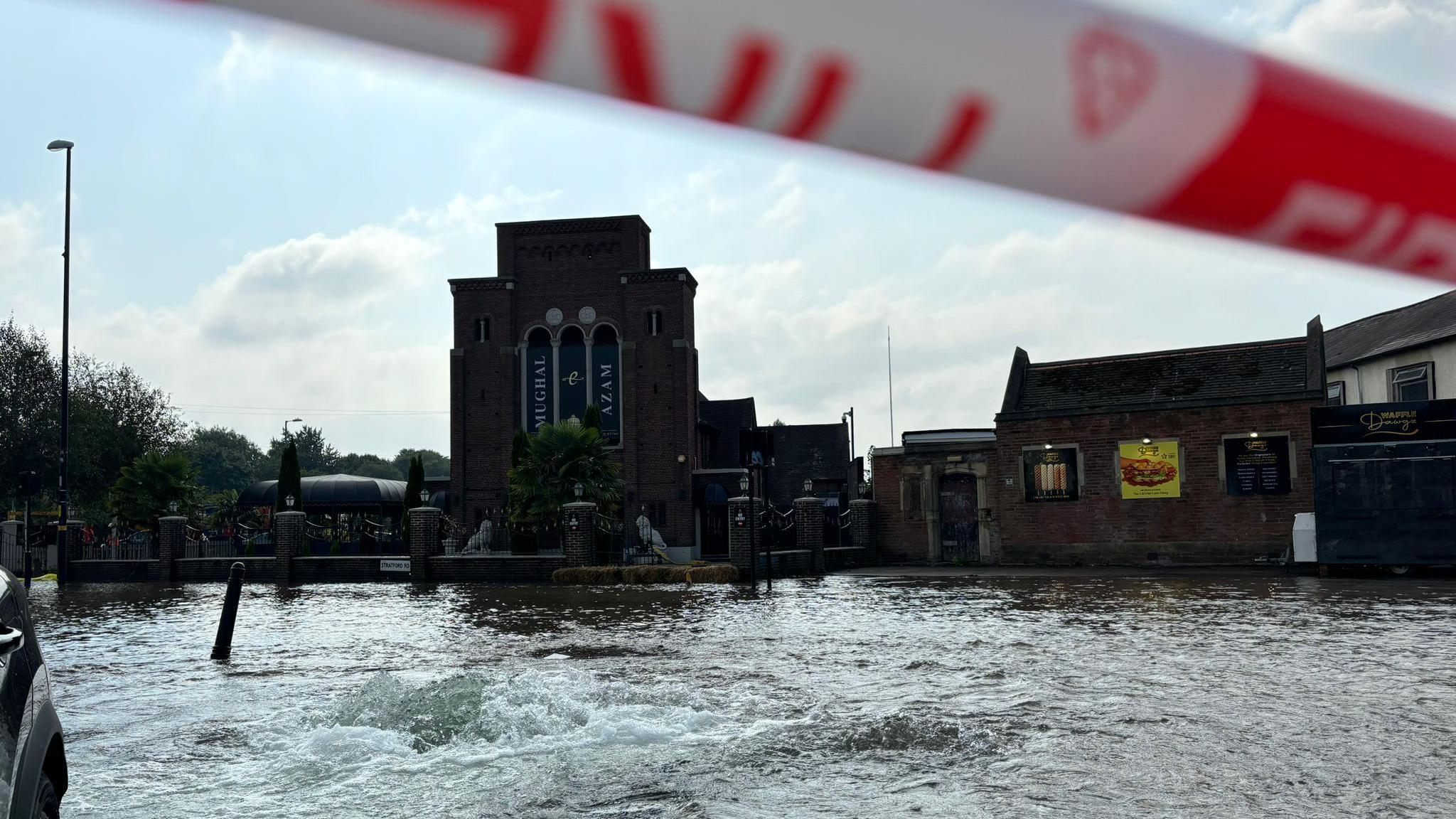 Flood-water in a street with a restaurant in the background. There is red and white "fire" tape at the top of the image