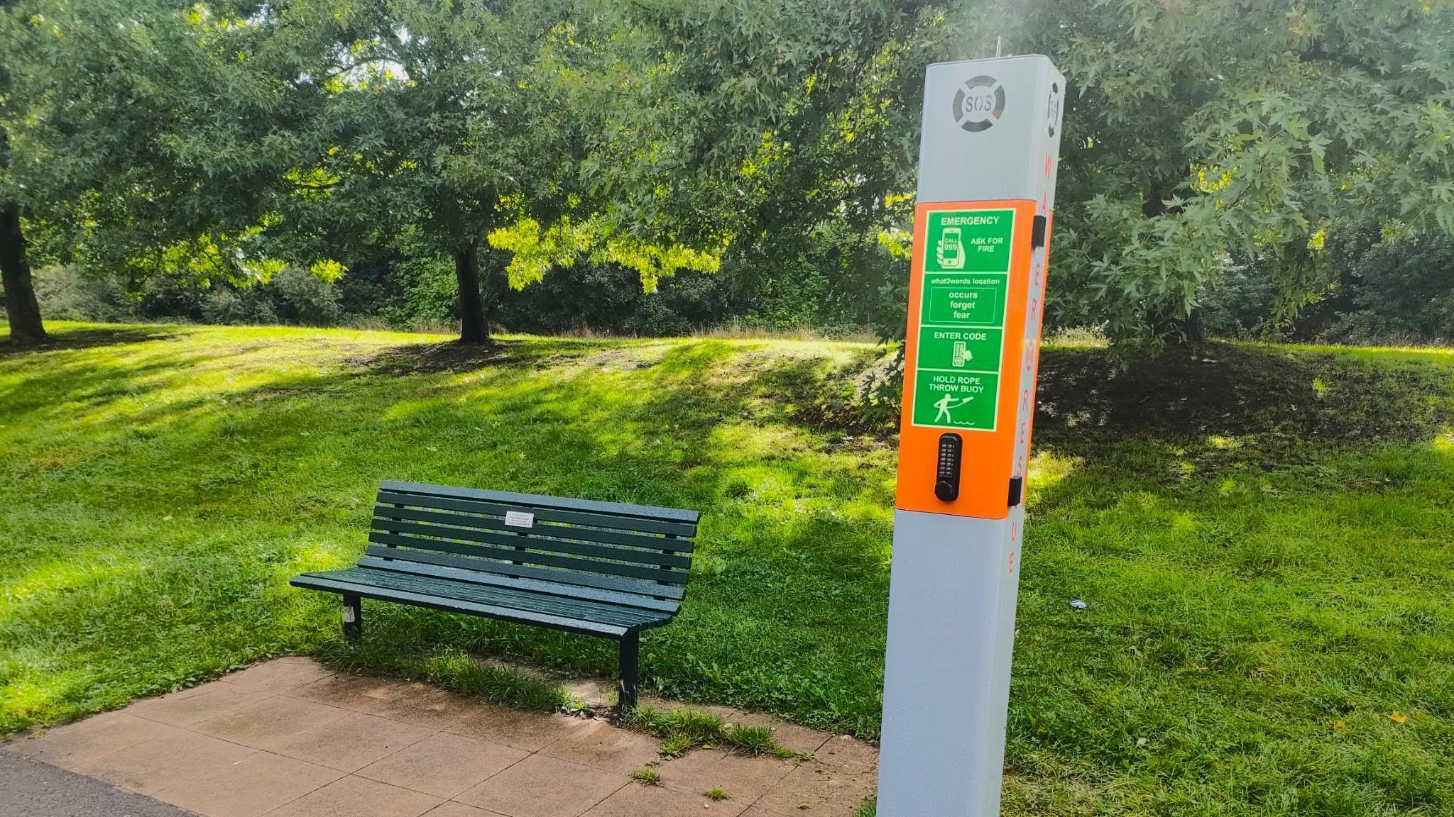 A green bench in a park next to a tall grey and orange cabinet with a green safety sticker attached which has instructions on. It has a keypad attached to it and a light box which has the letter SOS written over it.