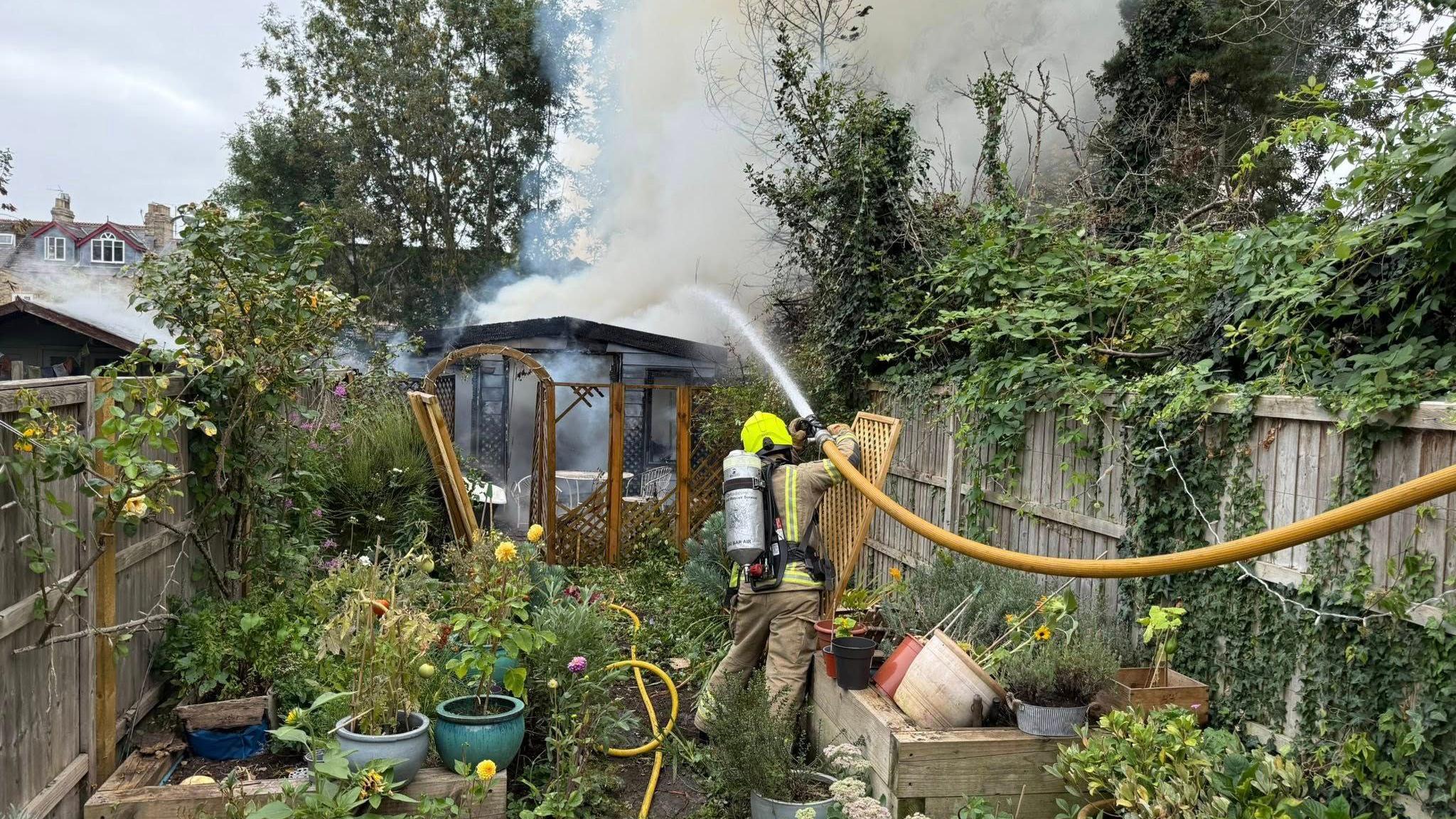 A firefighter is extinguishing the fire at a garden building with a large yellow hose. Smoke is hovering overhead. Smoke can also be seen coming from the roof of a neighbouring shed. The garden is full of pots of flowers and greenery. It is an overcast day.