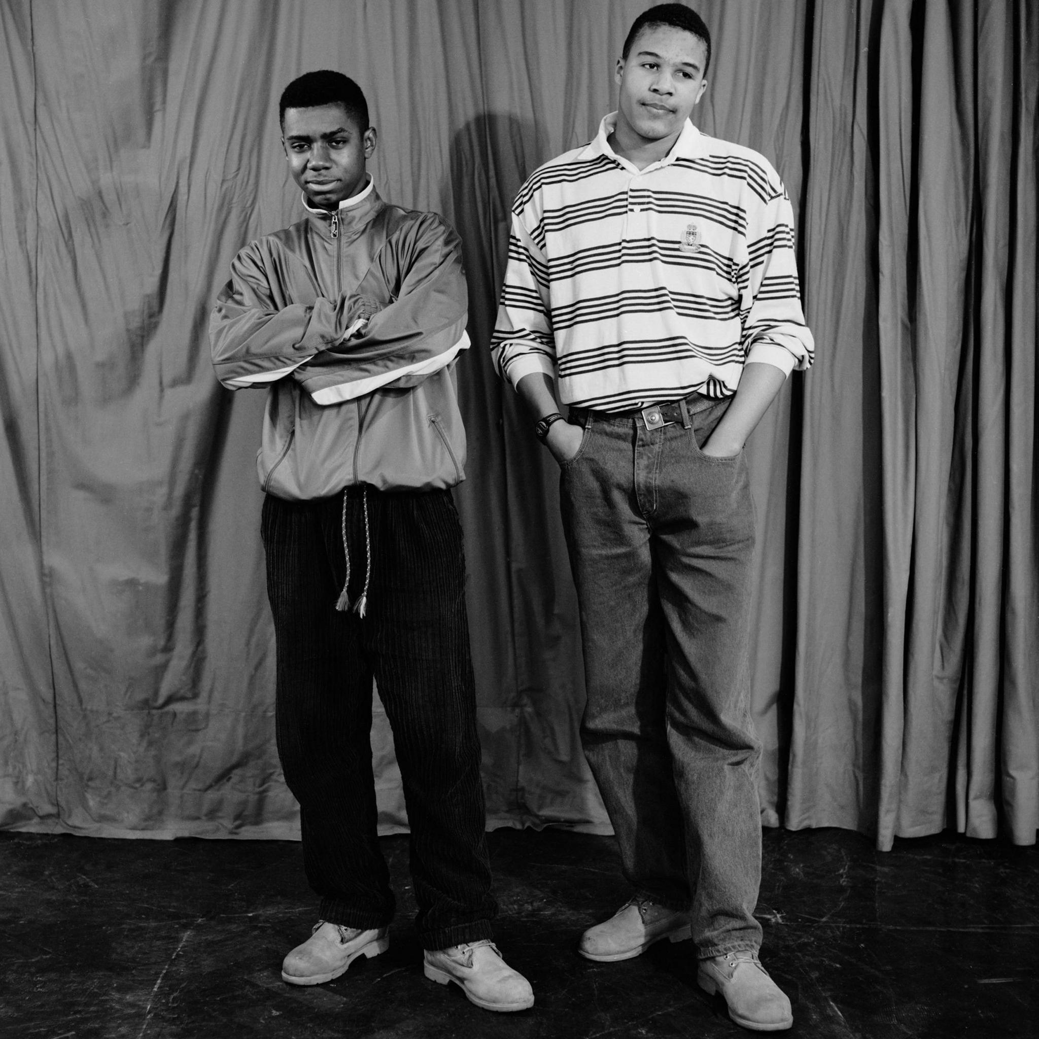 Boys from Tulse Hill School, 1990. Two young men stand in front of a curtain. One in corduroy trousers and a sports jacket Another in jeans and a striped top. Black and white photo.