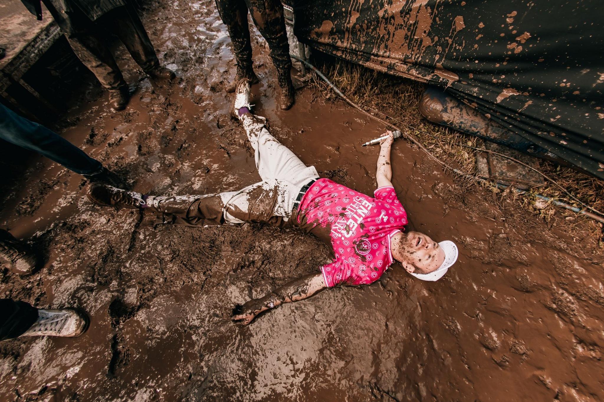Enter Shikari wearing a pink t-shirt and cream pants lies in a pool of mud while holding his mic in one hand