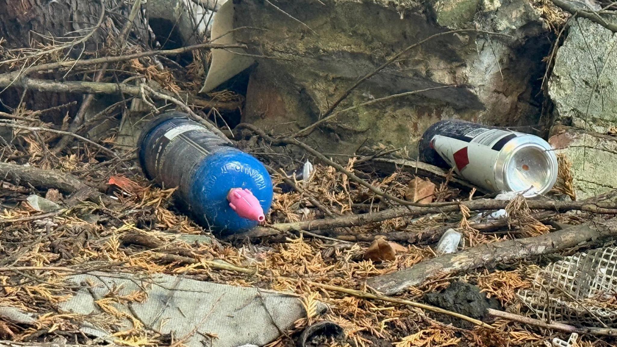 A nitrous oxide canister discarded on the ground, alongside an empty drinks can and other pieces of rubbish