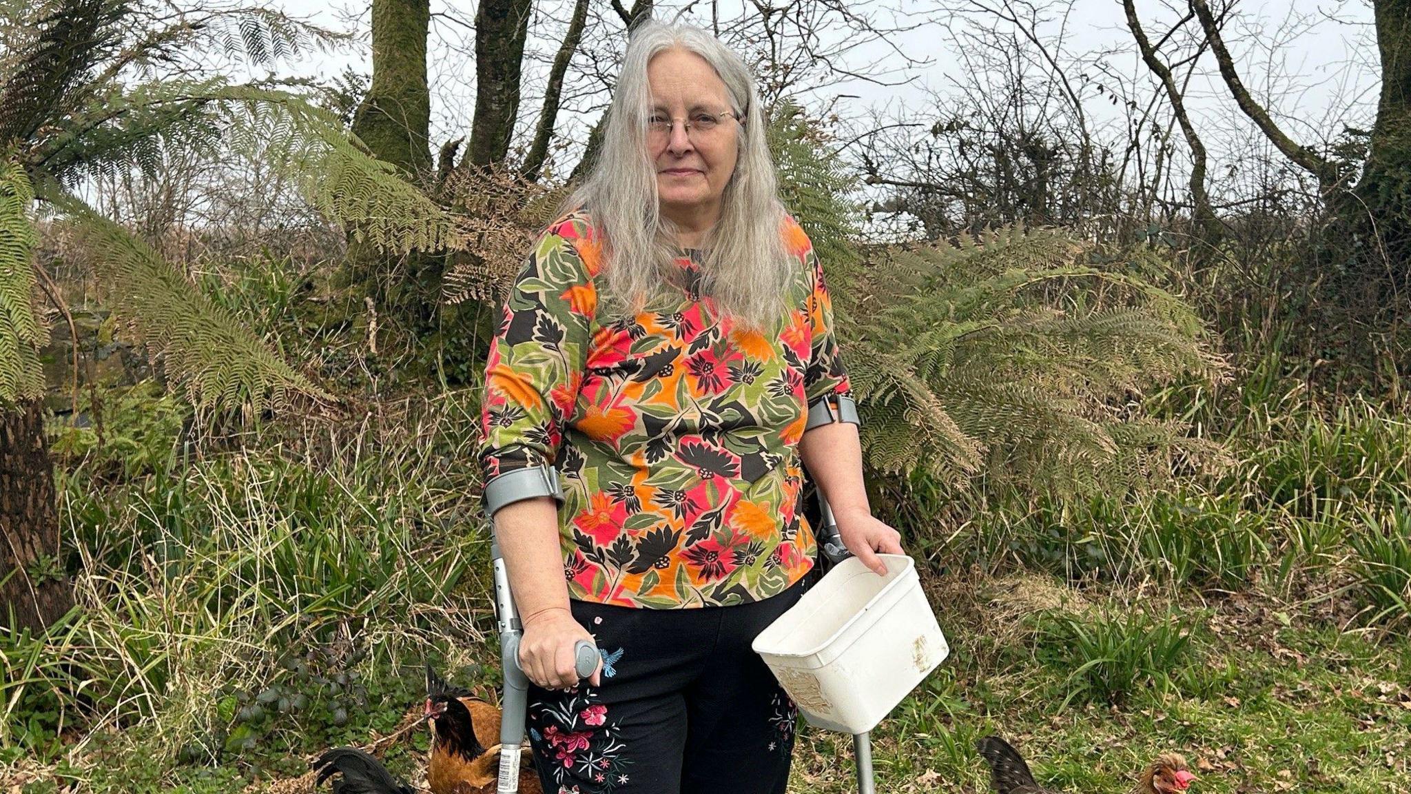 A woman with long grey hair in a brightly patterned top decorated with orange and green flowers. She is standing with crutches holding a white plastic pot with chicken feed in it