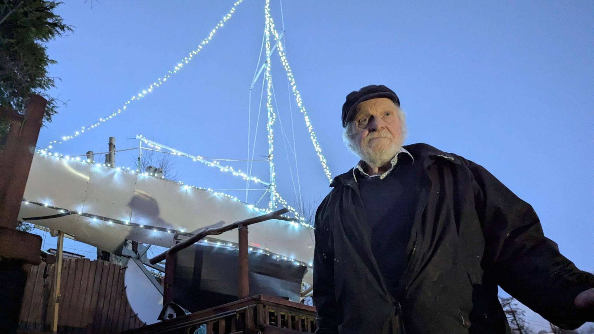 Ian standing in front of the 30ft yacht. He is wearing a sailor's cap and waterproof jacket with a knitted jumper underneath. He has a full grey beard.