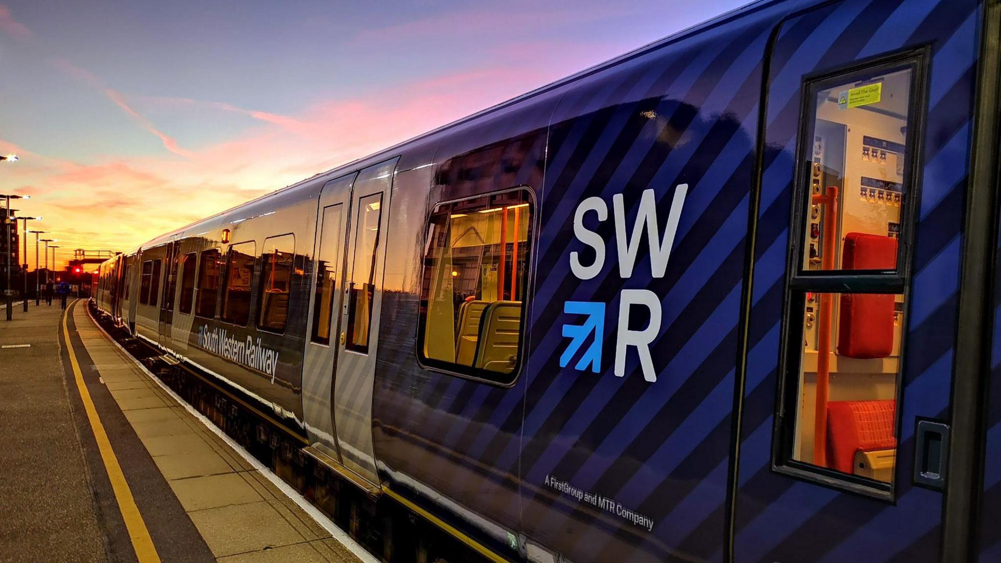 A blue liveried South Western Railway Class 450 train is in the platform at Basingstoke Station just before sundown.