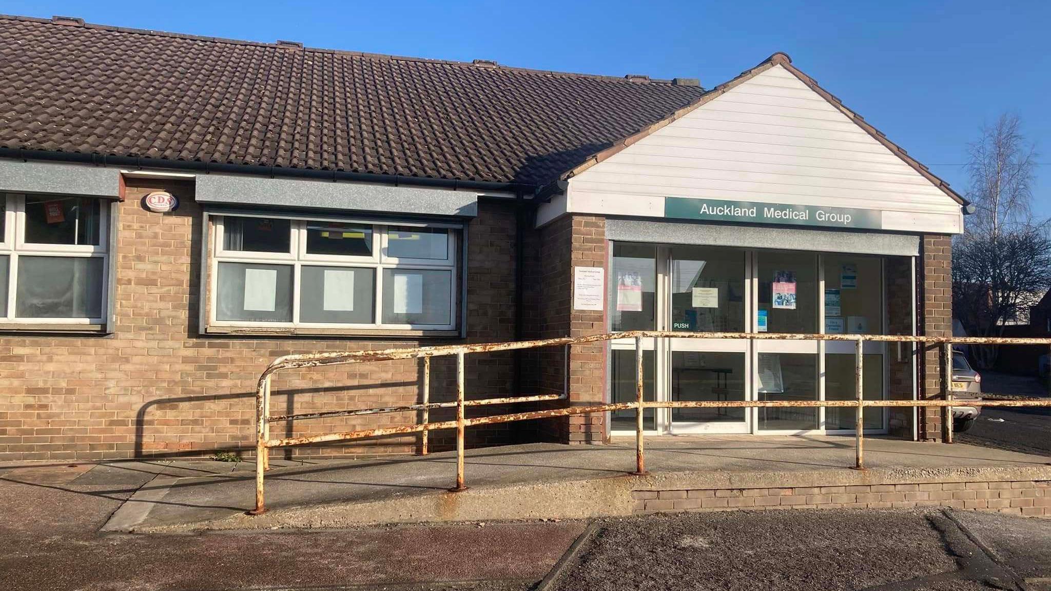 The front of the St Helen Auckland surgery has a rusty rail on either side of a ramp leading up toe a white door. It's a brick built building. 