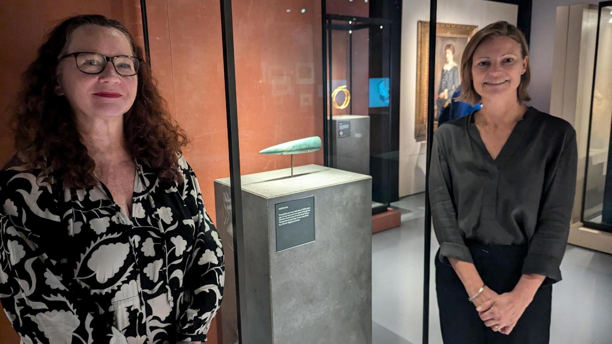 Two women stand next to a museum exhibit of a light blue tooth-shaped object.