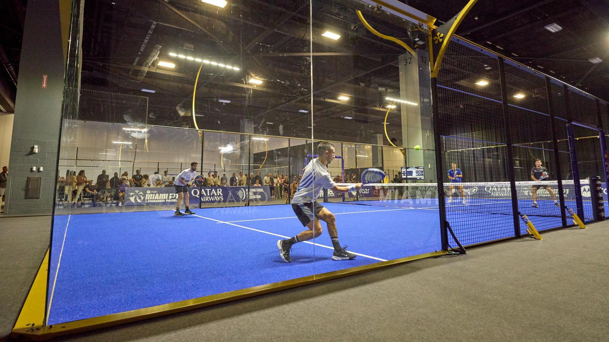 A padel court, with the view coming from outside the court. A match is ongoing on the blue turf.