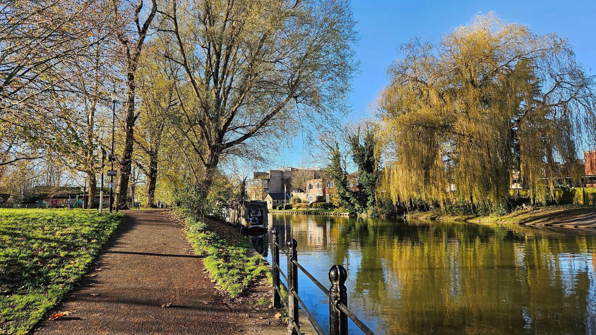 A river running to the right of a path with trees and buildings in the background under a blue sky