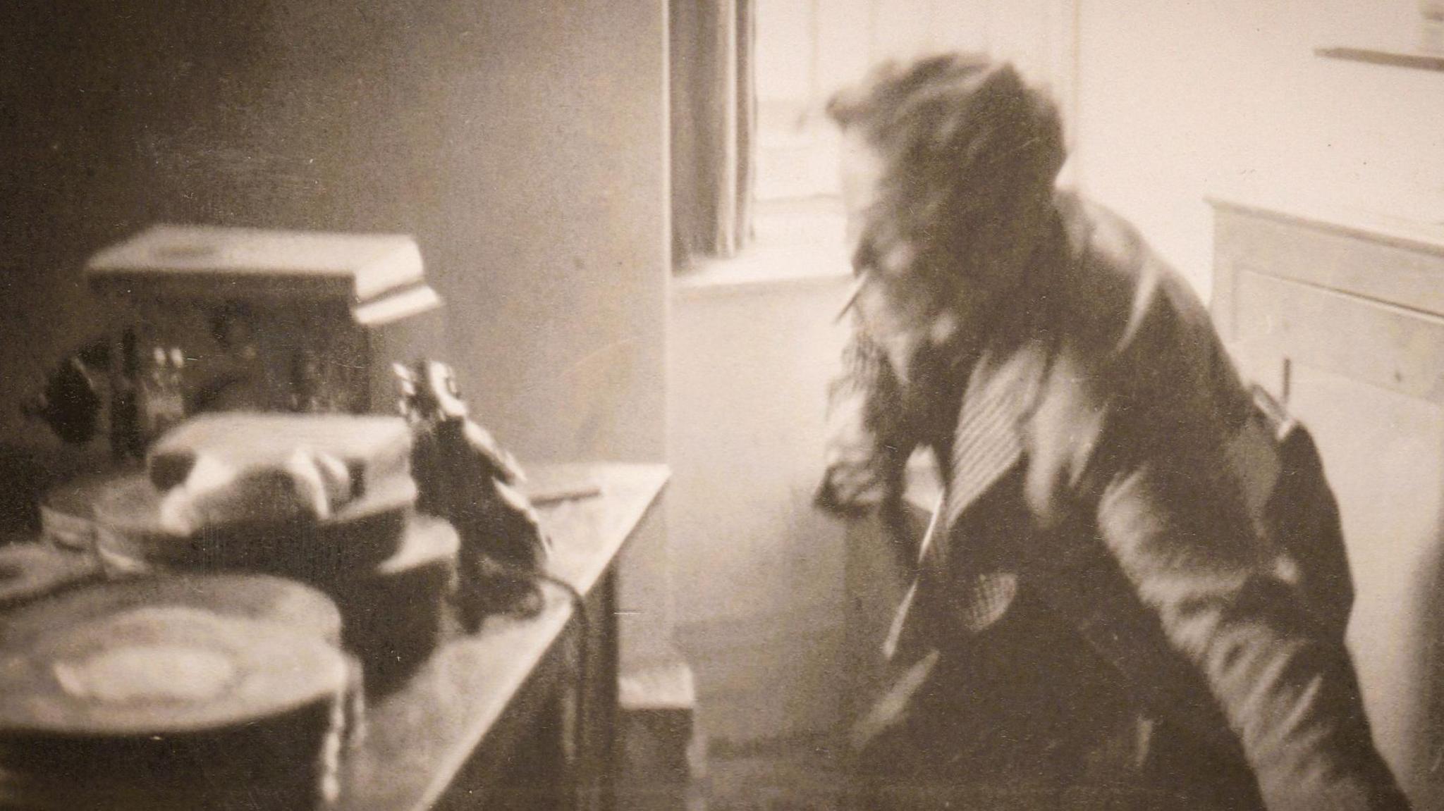 Dylan Thomas in a black and white photo standing beside the same window but is crouching with the chair legs still in one hand. A table beside him has audio recording equipment from the era and film cans stacked up.