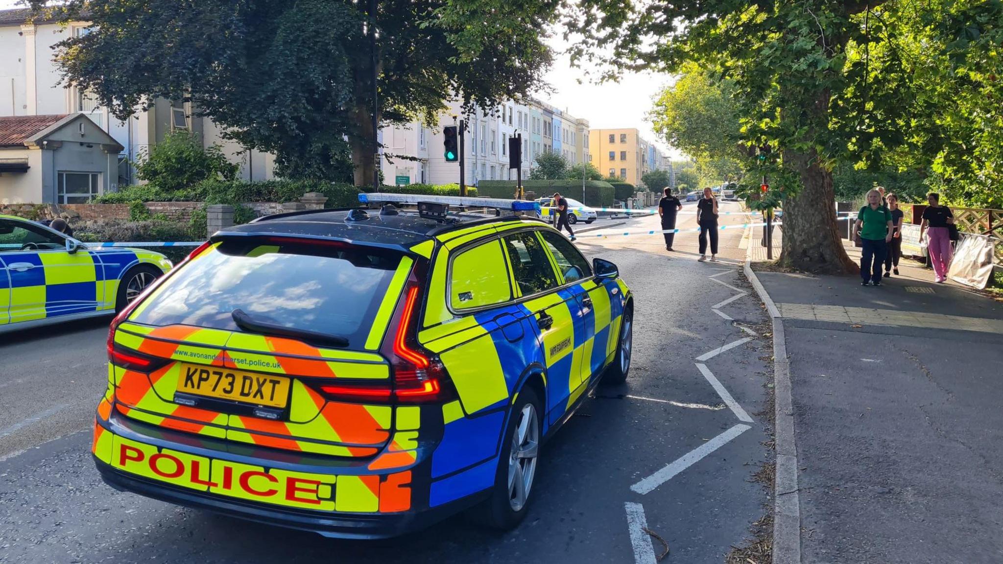 Two police cars in front of a cordon on Coronation Road. Police tape can be seen across the road, with police officers visible in the distance. 