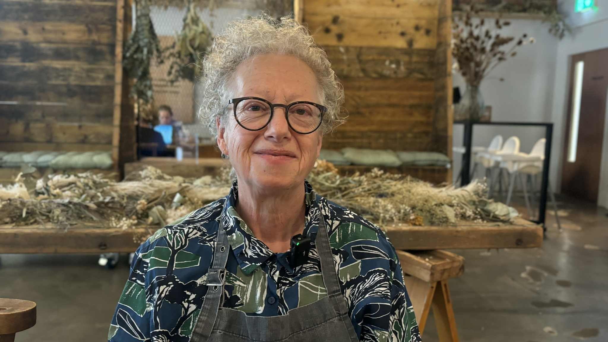 Jill Barnett looks at the camera. She has short grey curly hair and black-rimmed glasses and is wearing a blue and green patterned shirt beneath a grey apron.