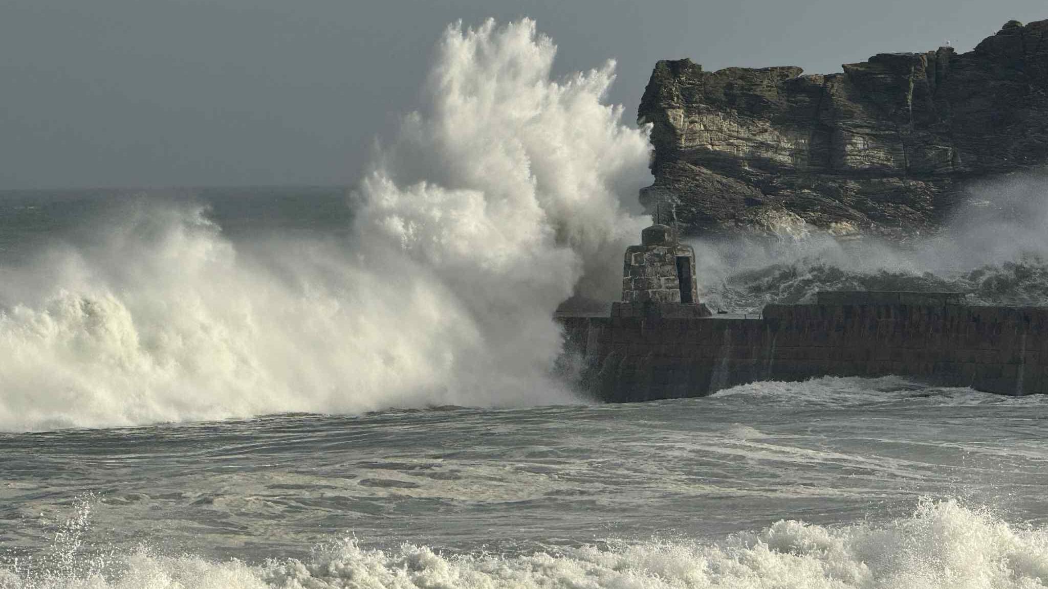An enormous wave is crashing on a harbour wall. The breaking wave is creating a huge white wall of water surging to the shore. The wave is lit by the sunshine and the headland is rocky and steep.