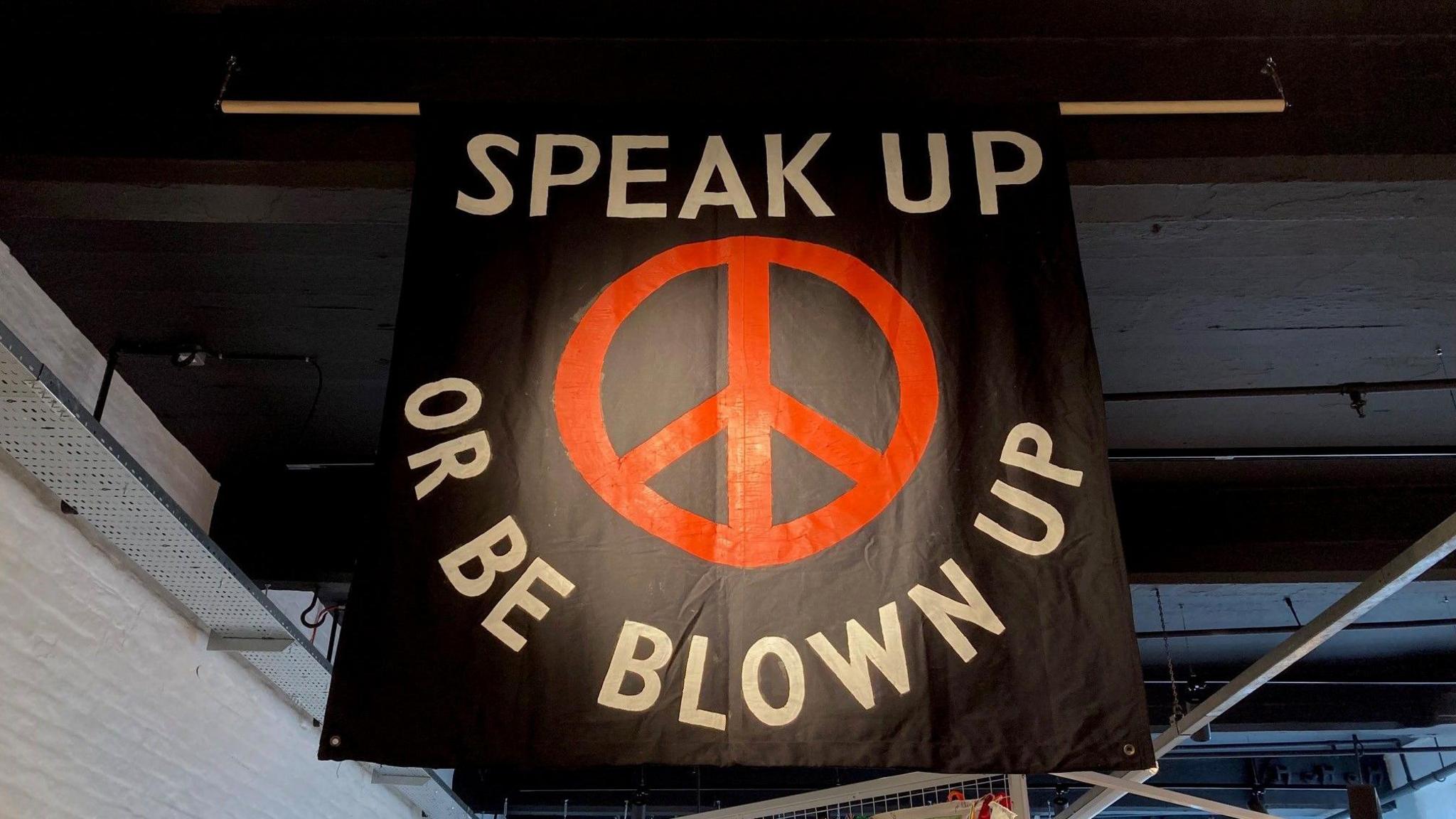 A peace banner hanging from the ceiling with the words 'Speak up or be blown up' spelled out in white lettering on a black background circling around a red peace sign. 