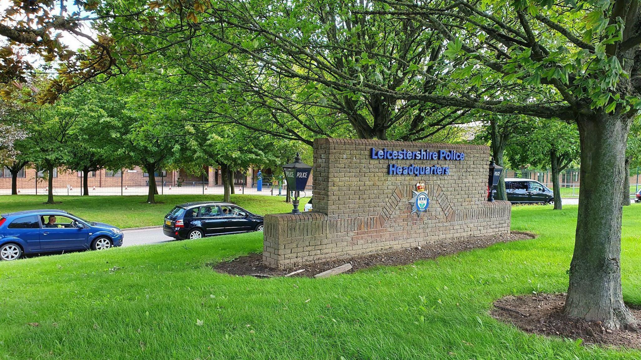 Leicestershire Police Headquarters. A brick call can be seen with blue lettering which reads "Leicestershire Police Headquarters". It is surrounded by trees.