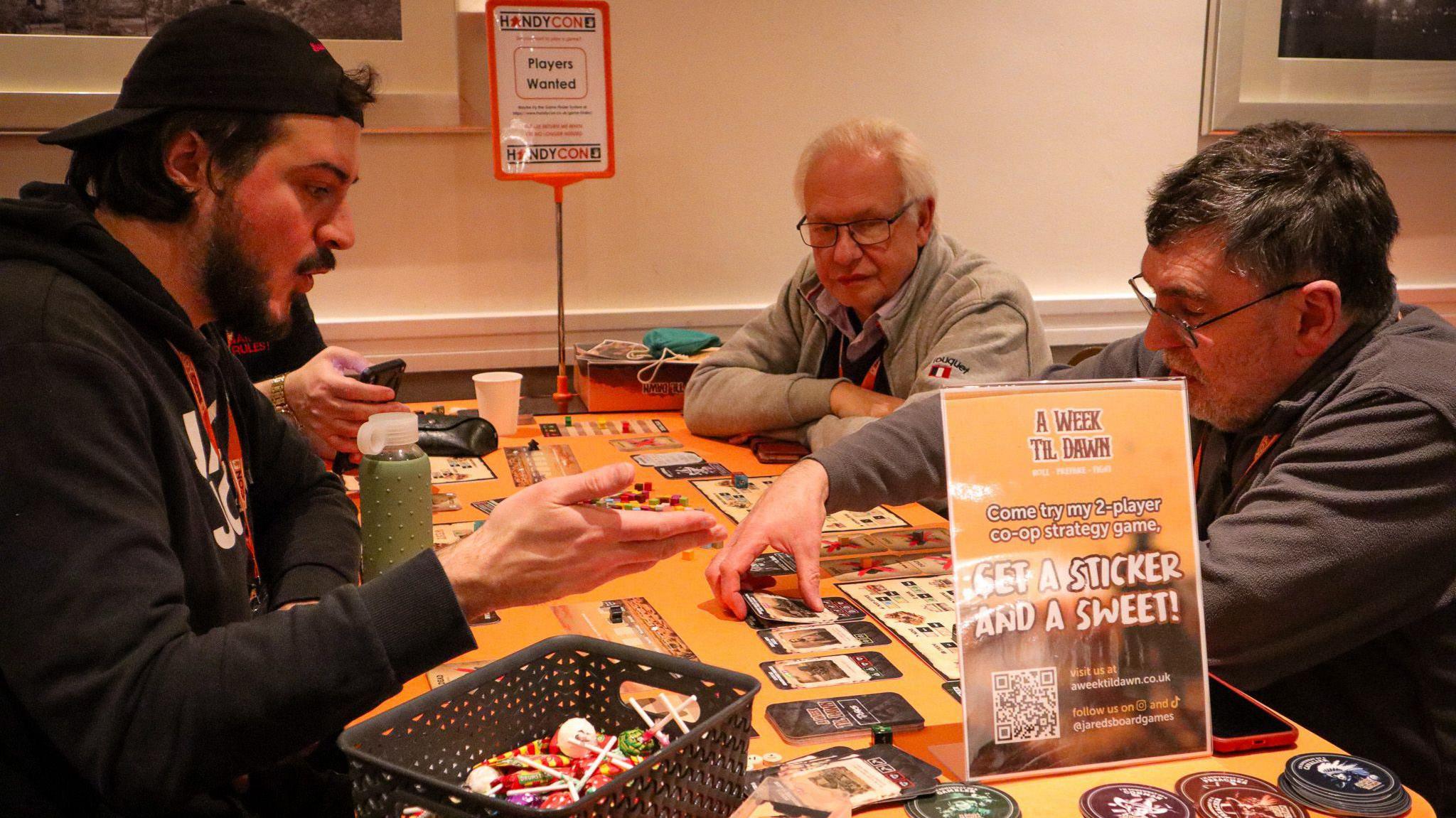 A group of men of various ages are sat at a table covered in cards and other board game paraphernalia