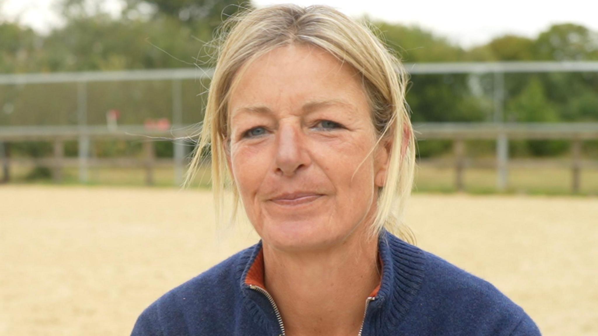 Zannie Howard smiles as she sits inside an outdoor equestrian school. She has blonde hair that has been tied back. She wears a navy knitted jumper.