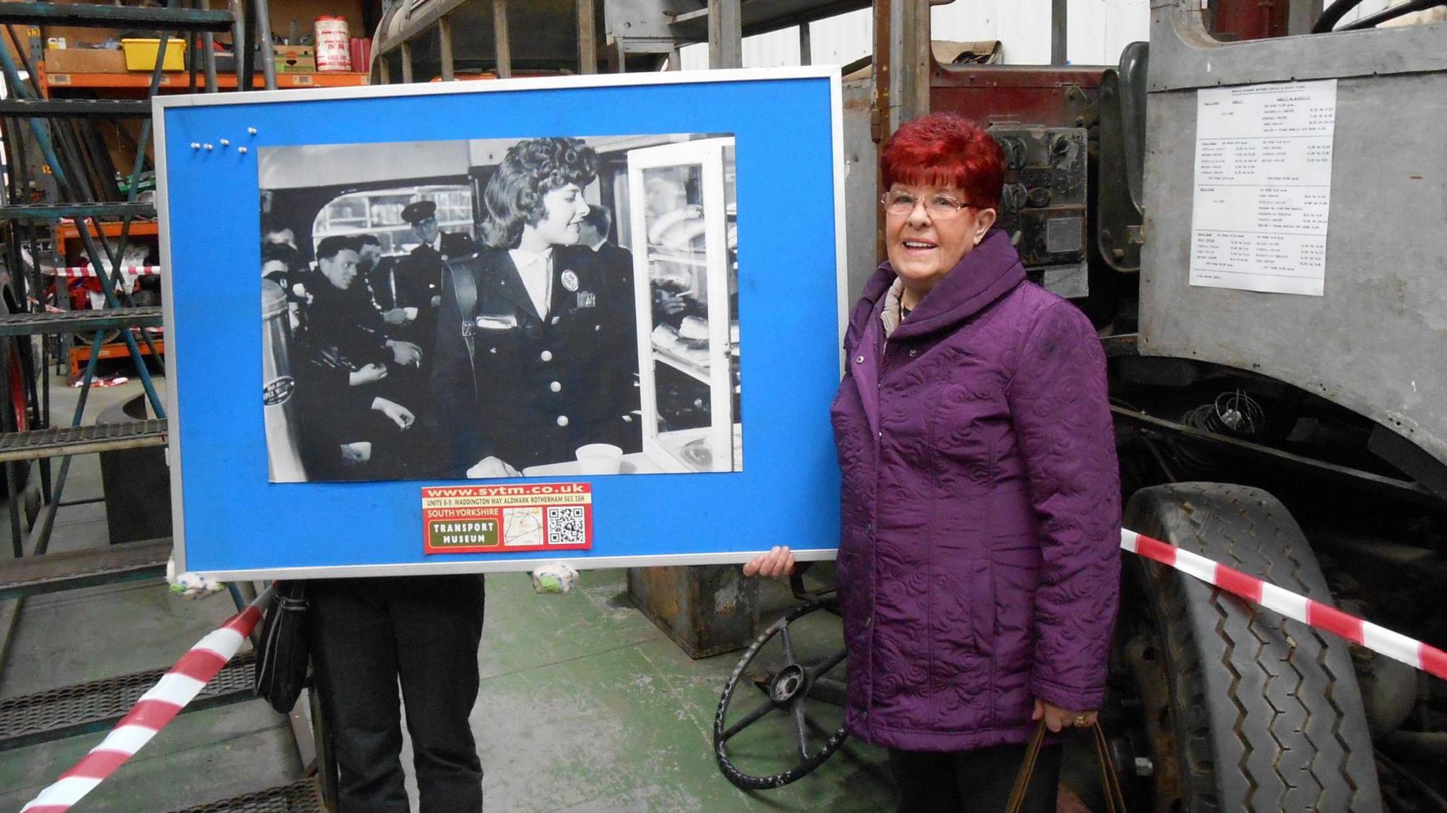 A woman wearing a purple coat, Julie Askew, stands next to a large print of a photograph of her mum wearing a bus conductor's uniform