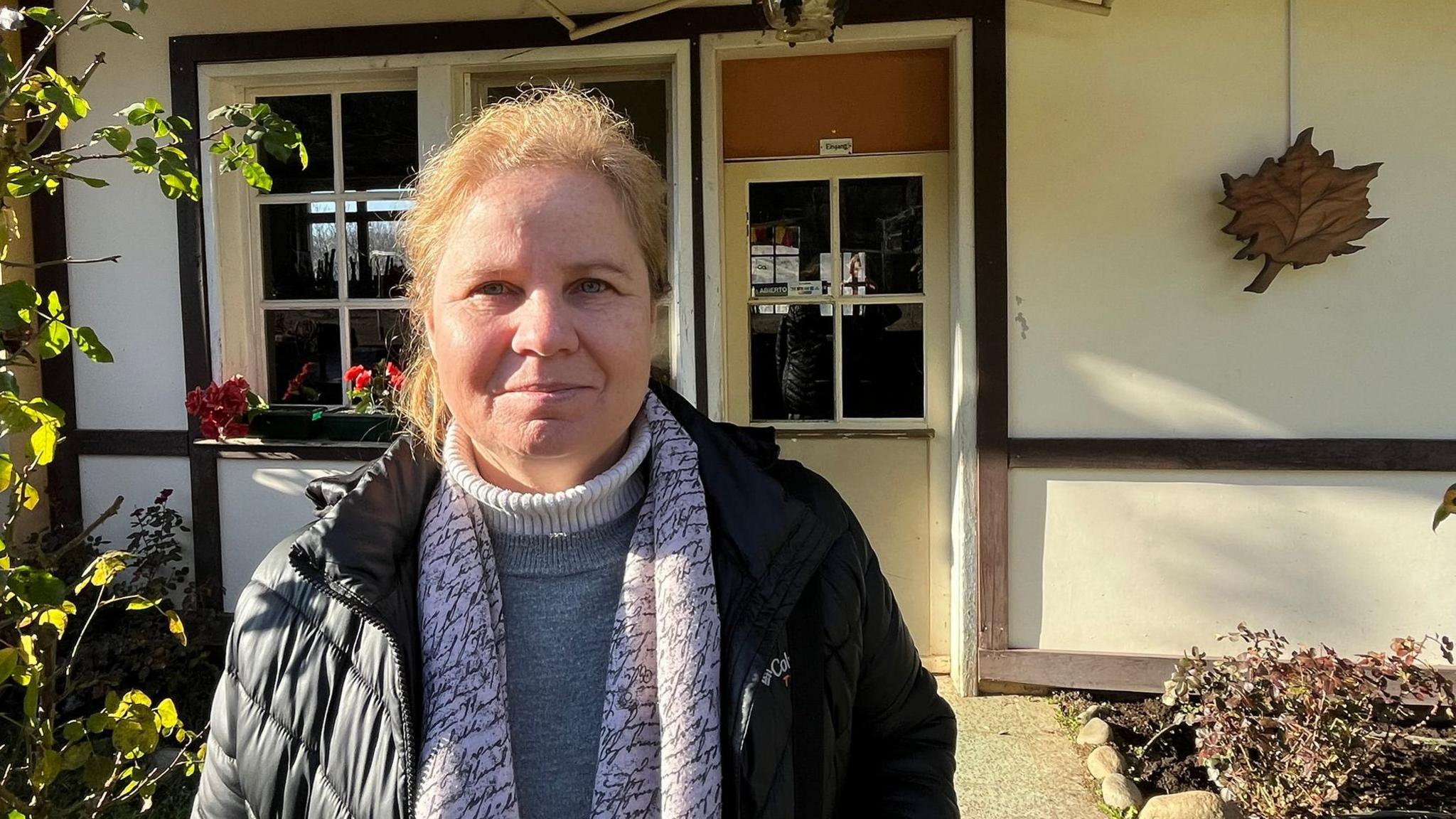Dorothee Munch, wearing a black puffer jacket and a white scarf, stands in front of one of the houses in Villa Baviera. 