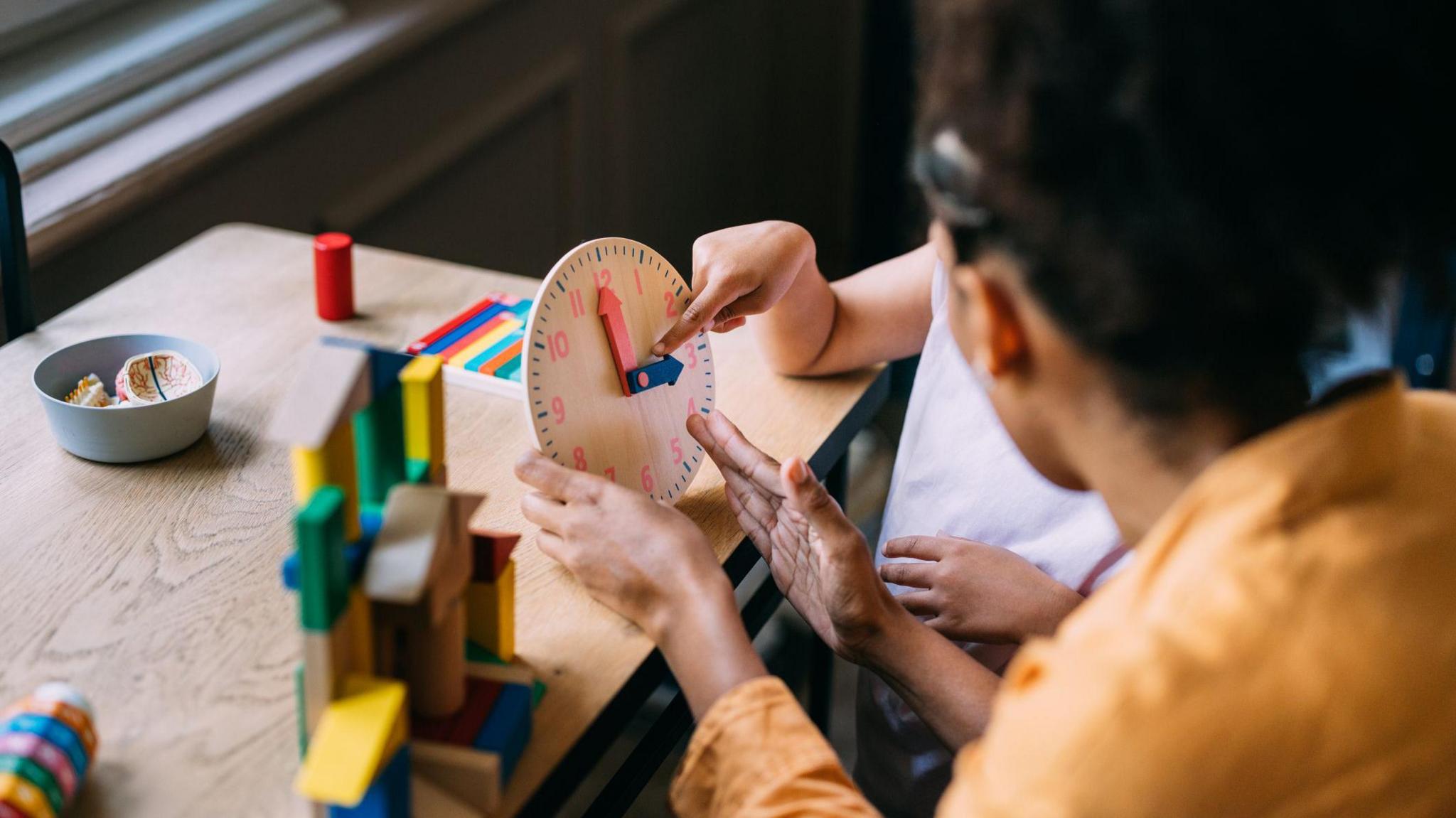 An adult teaches a child how to tell the time using a small clock
