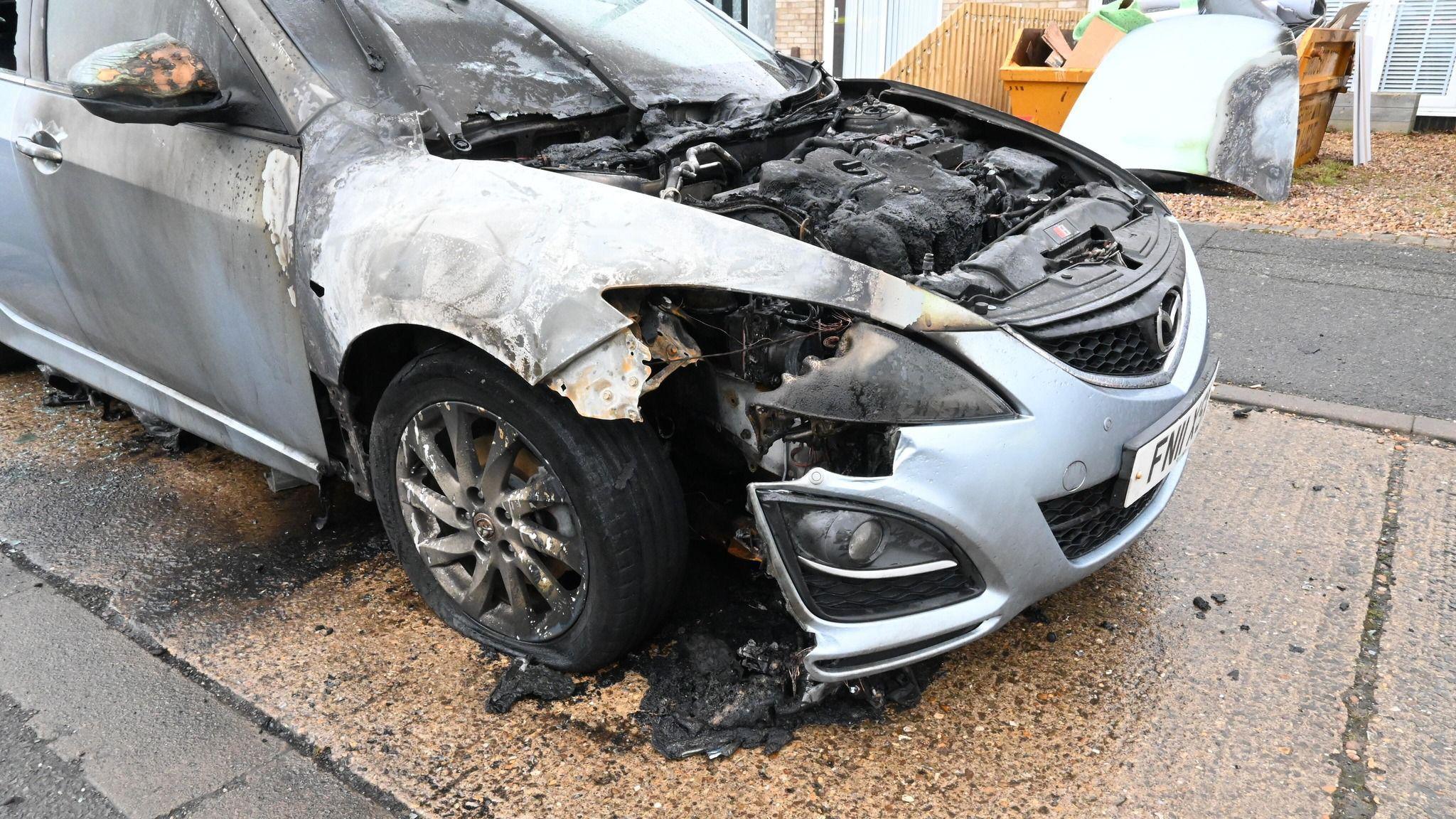 The front of a car badly affected by a fire. It is a silver Mazda but is badly charred. It is parked on a road or driveway.