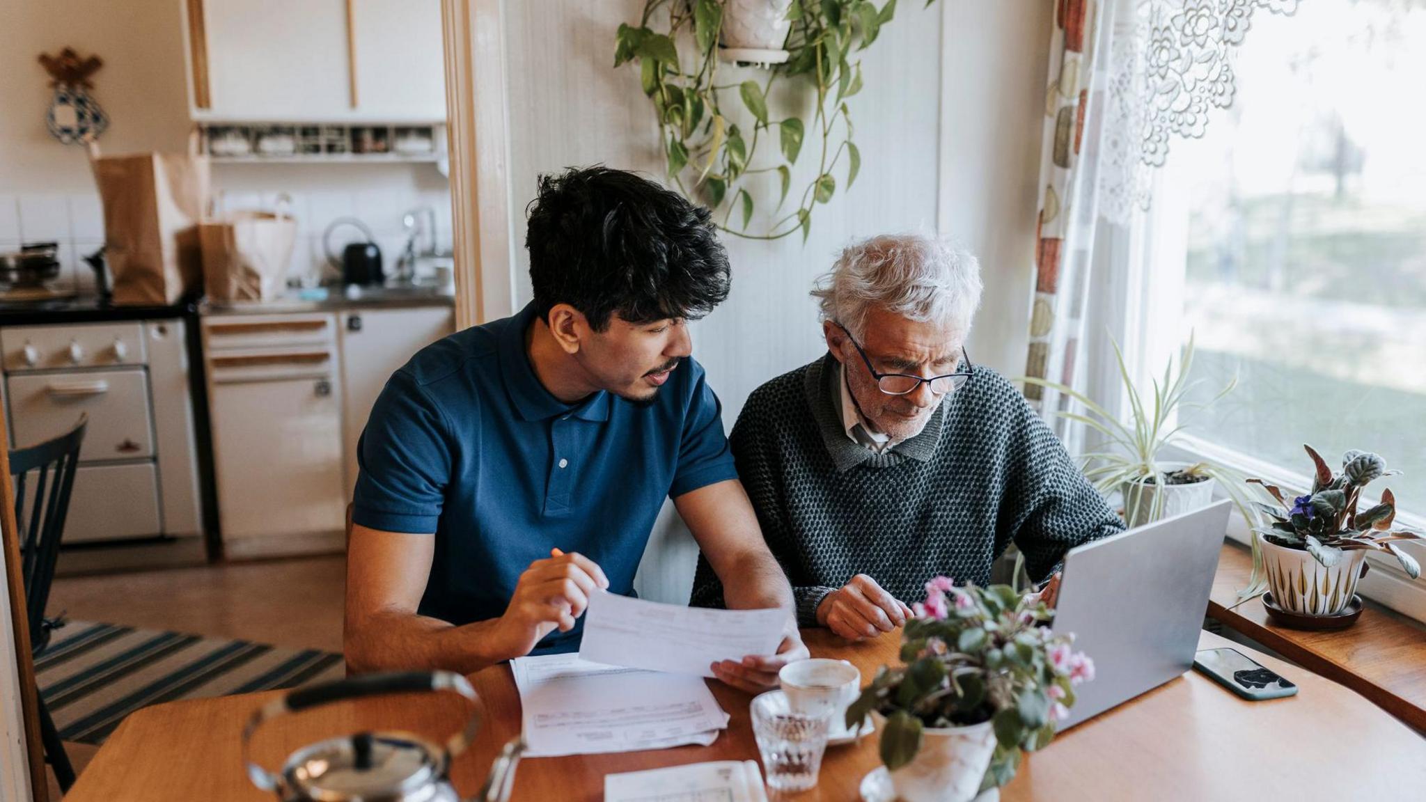 A young man sits next to an older man at a kitchen table as they look at a laptop and go through paperwork. 