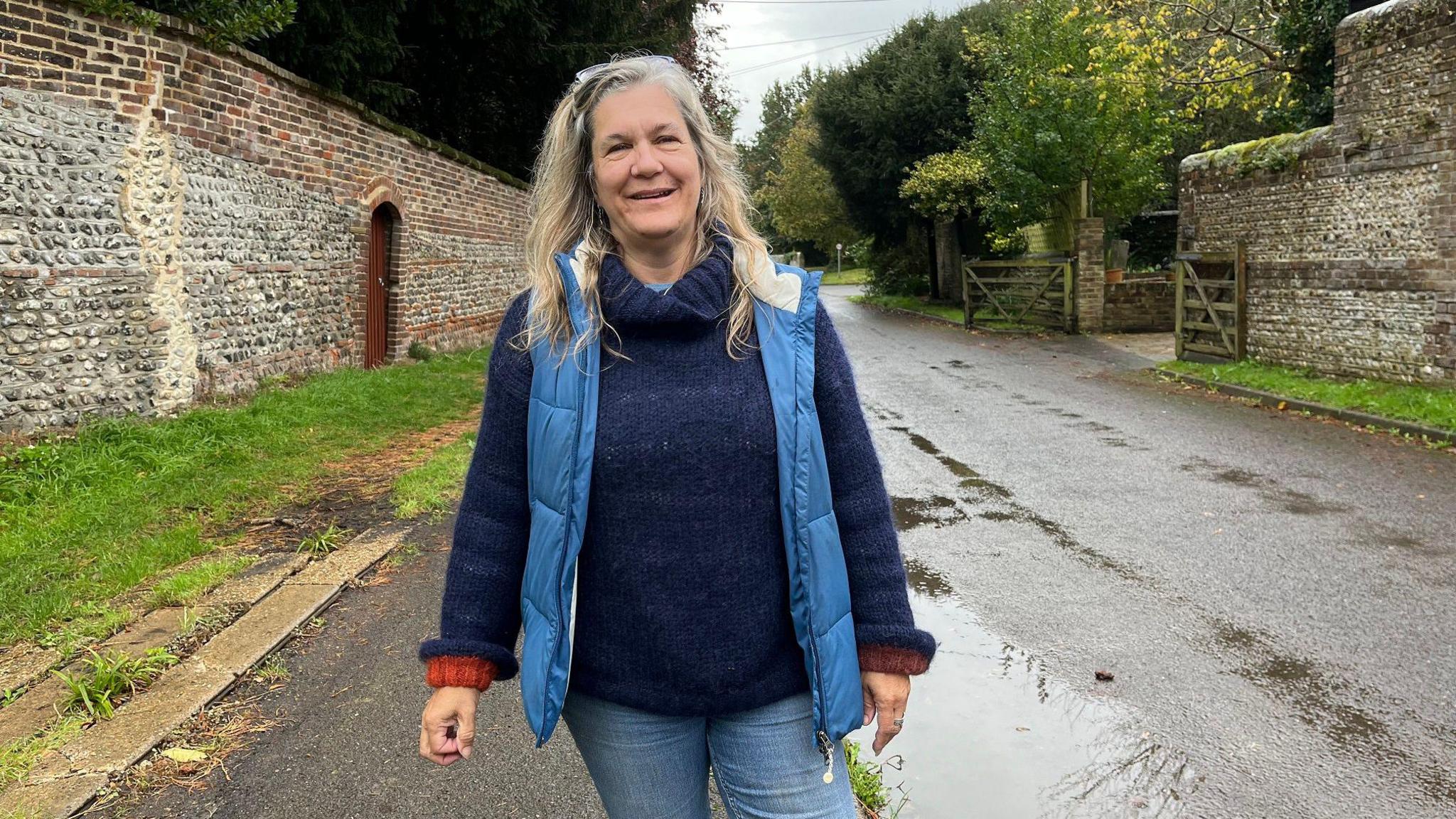 Claire Messenger wearing a padded blue gilet and standing in a rural-looking quiet road with brick and stone walls