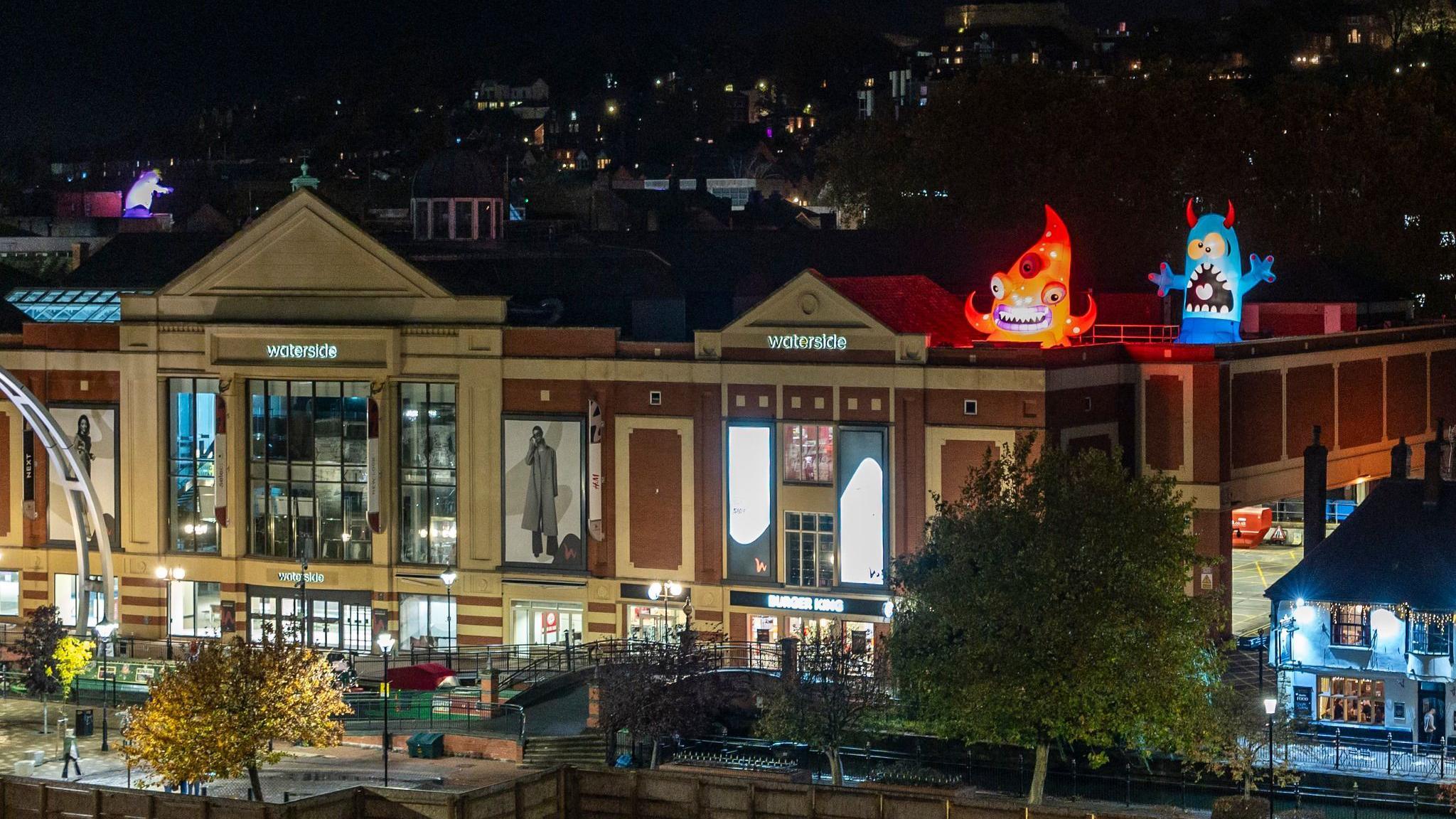 A shopping centre building can be seen at night time which reads 'waterside' on the front, next to a pub. Lights from other buildings can be seen in the distance. An orange inflatable monster with a purple smith and a blue monster with red horns are lit-up on top of the building.