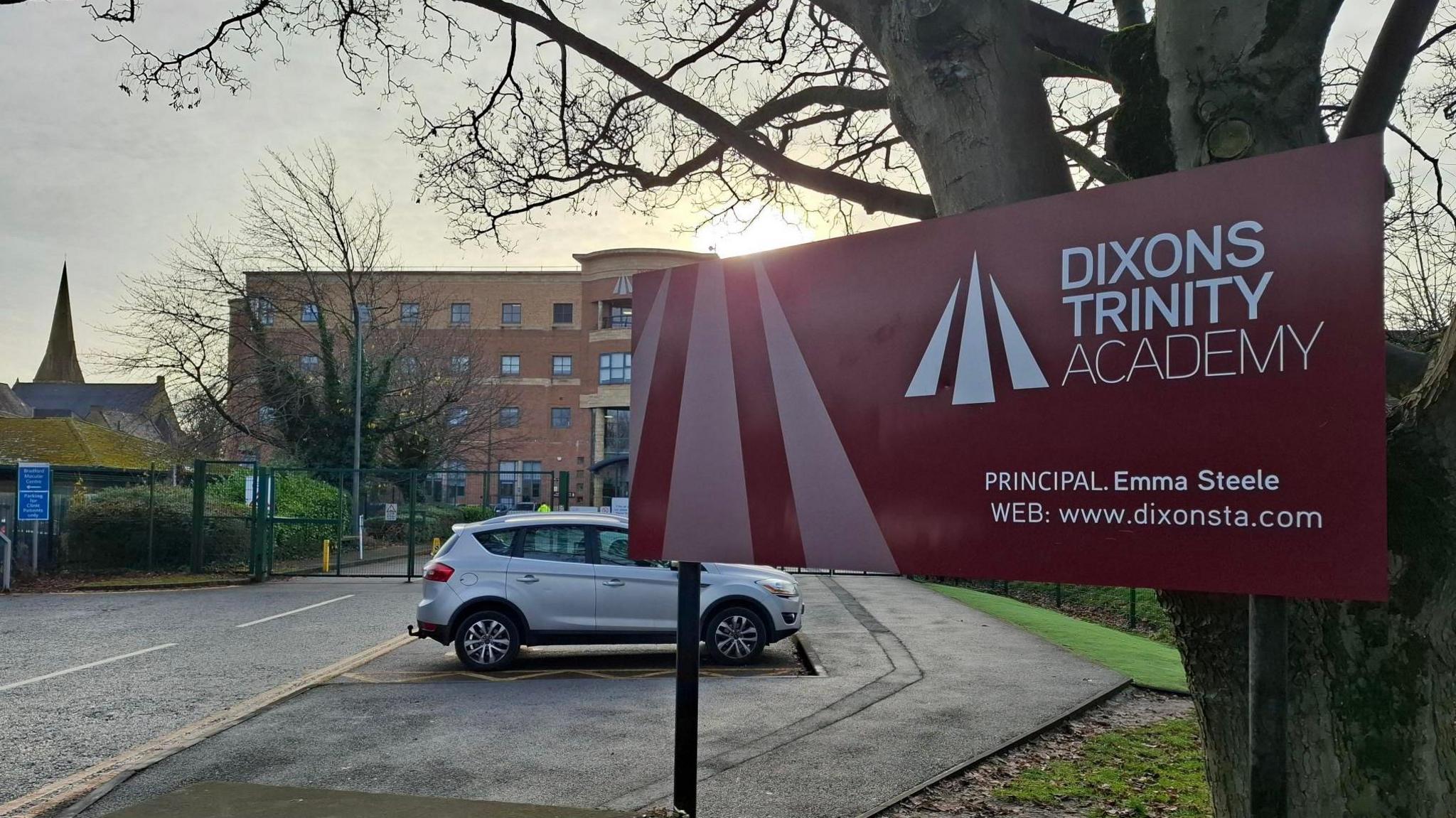 A red sign reading Dixons Trinity Academy Principal Emma Steele Web www.dixonsta.com in front of a car park and a four storey building. A silver car is parked behind the sign.