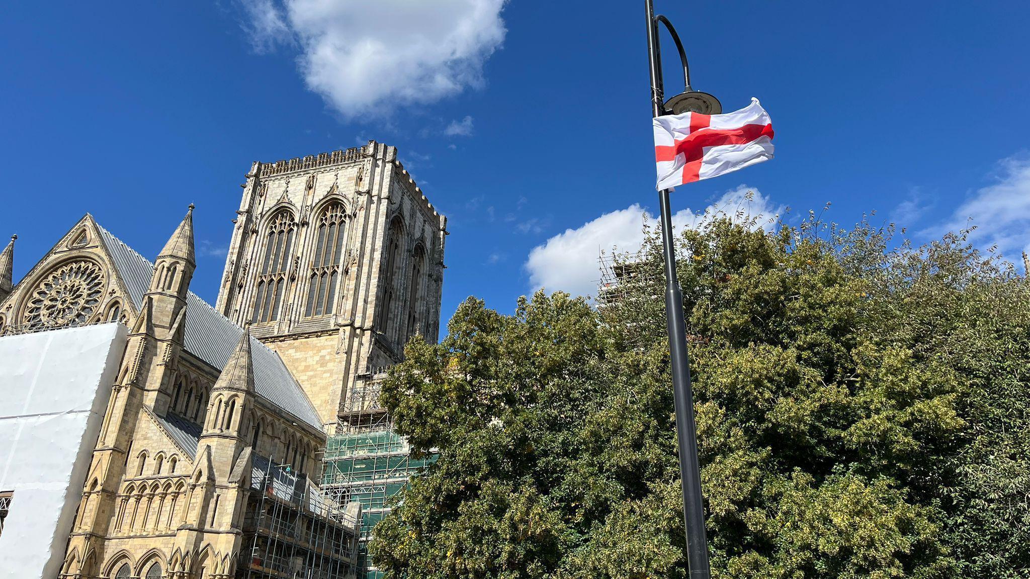 An England flag sits on a lamppost against the backdrop of York Minster. Some trees are also in the background.