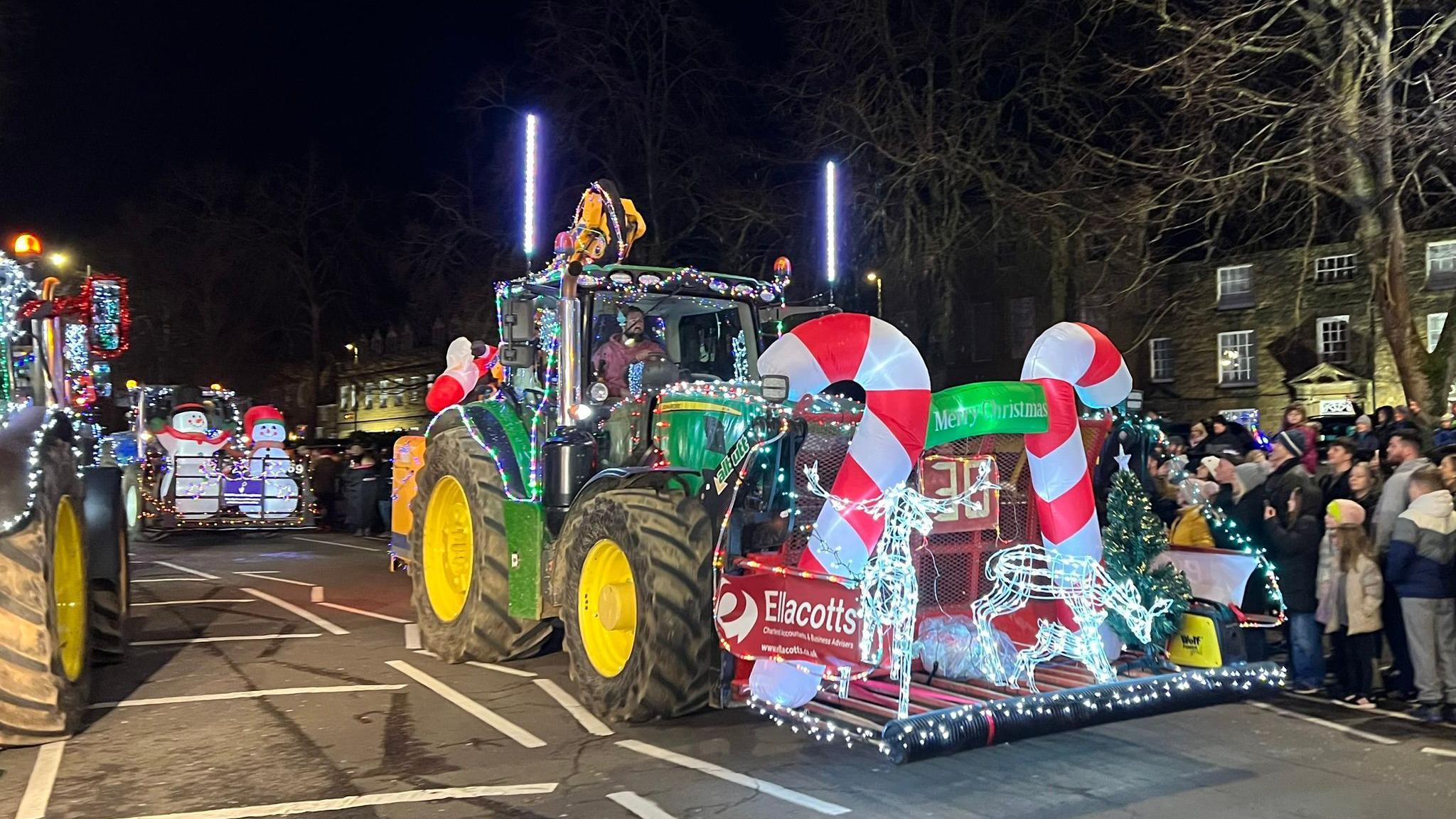 A large green tractor decorated with lights and large candy canes at the front. There are other decorated tractors around and crowds on the side of the street.