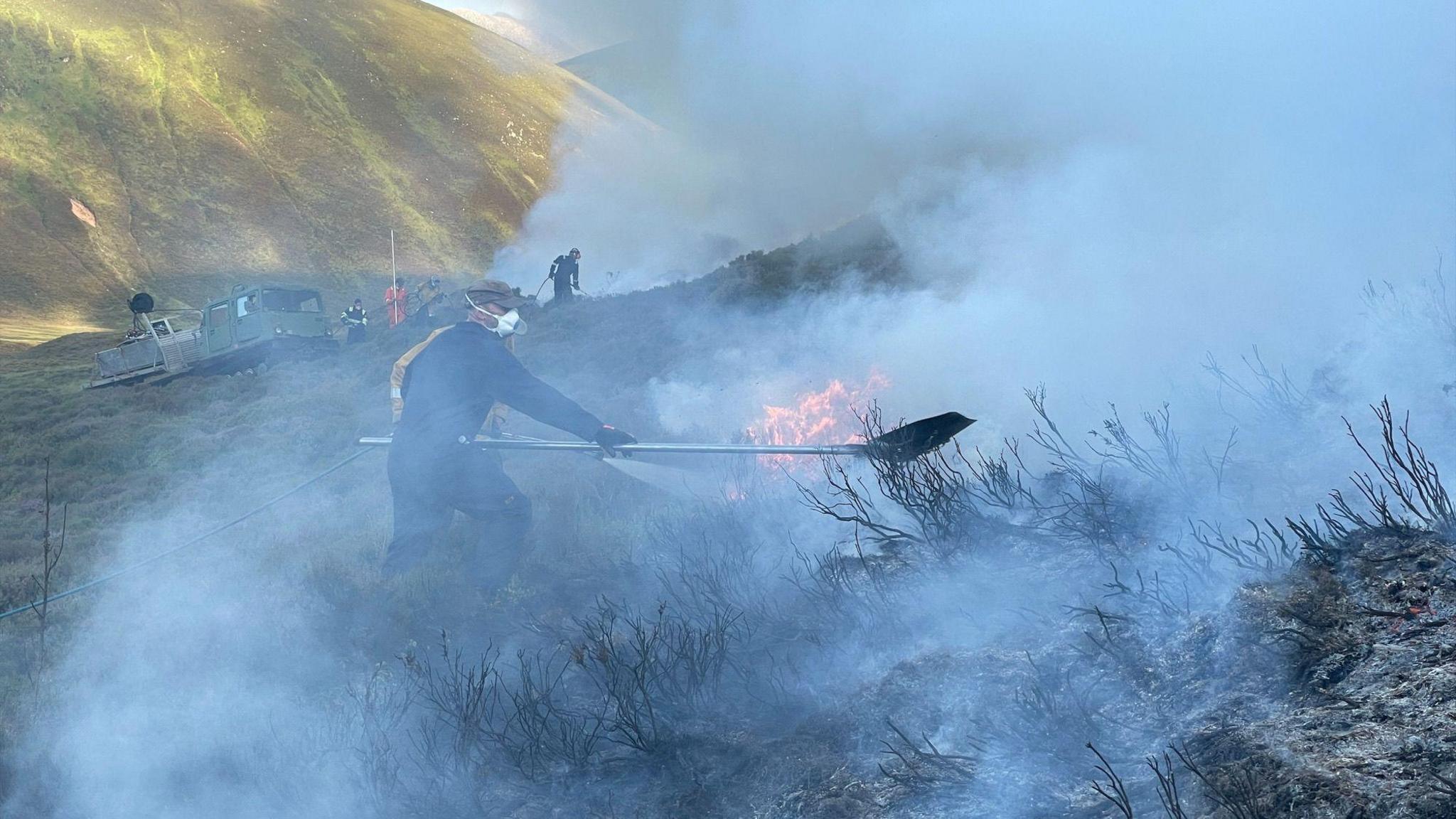 Daoine a' strì le teine fiadhaich sa Mhonadh Ruadh le masgaichean orra agus iad a' bualadh air an talamh le spaidean mora
