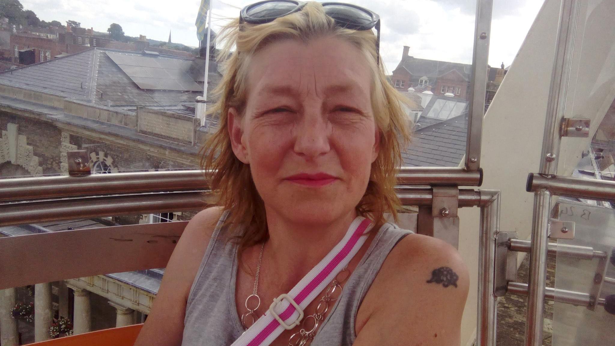 Dawn Sturgess looking at the camera. She has blonde hair and is wearing a grey vest top. She has sunglasses on her head and the strap of a bag over her shoulder is visible and is wearing a necklace.