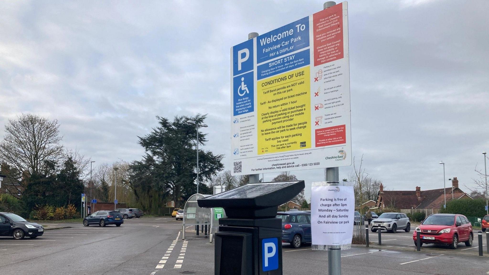 A view of one of the new parking machines with signs about parking charges. The machine is in a car park with a few cars behind it, red, blue and grey in colour. The machine has a solar panel on top, pointed at the sky and the big metal panel on a pole behind it has "P - Welcome To" at the top and then in smaller, hard to read text, the details of the parking charges and restrictions.