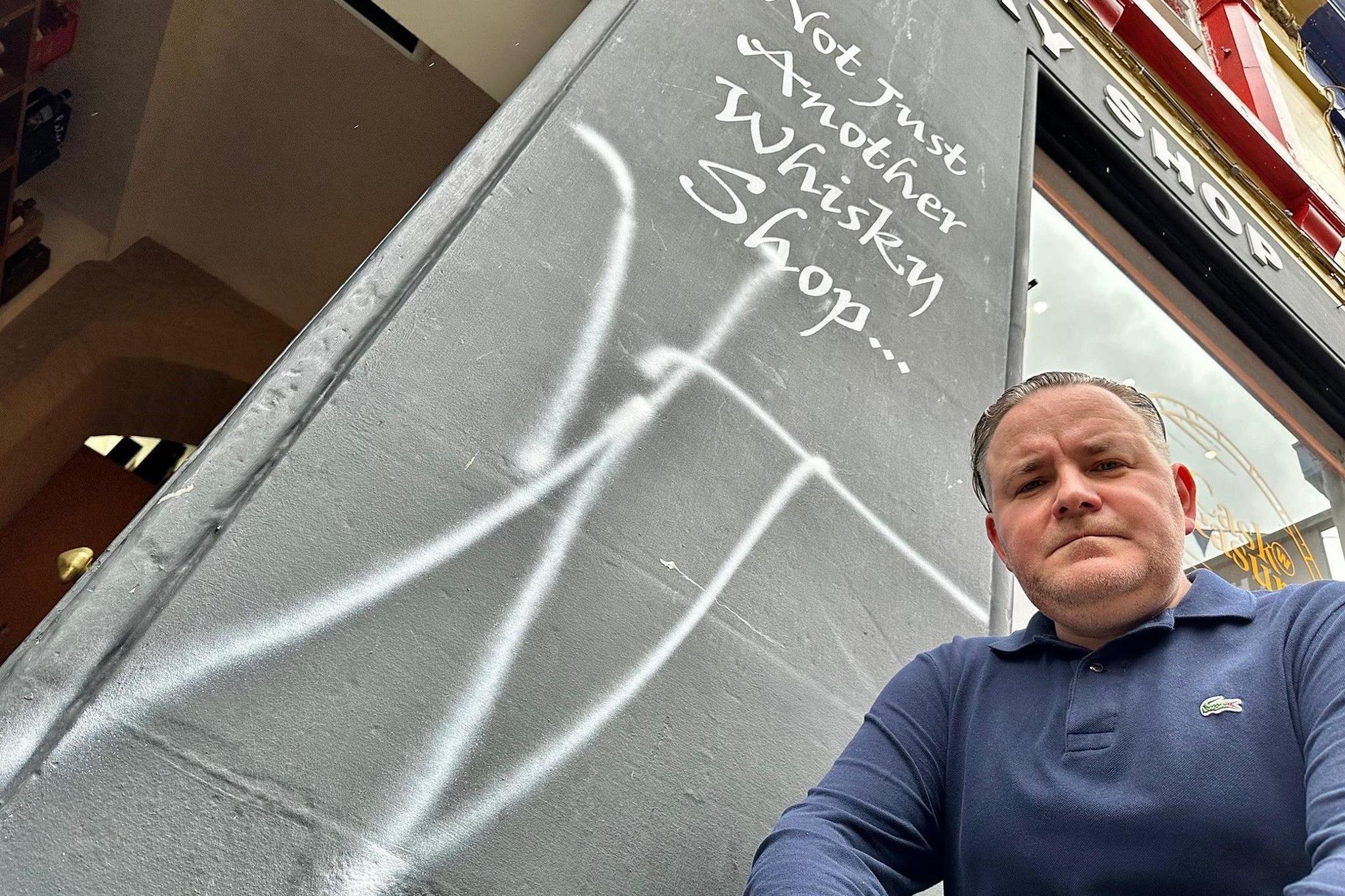 Andrew is looking down at the camera. He is wearing a blue top. There is white graffiti beside him on a shop.