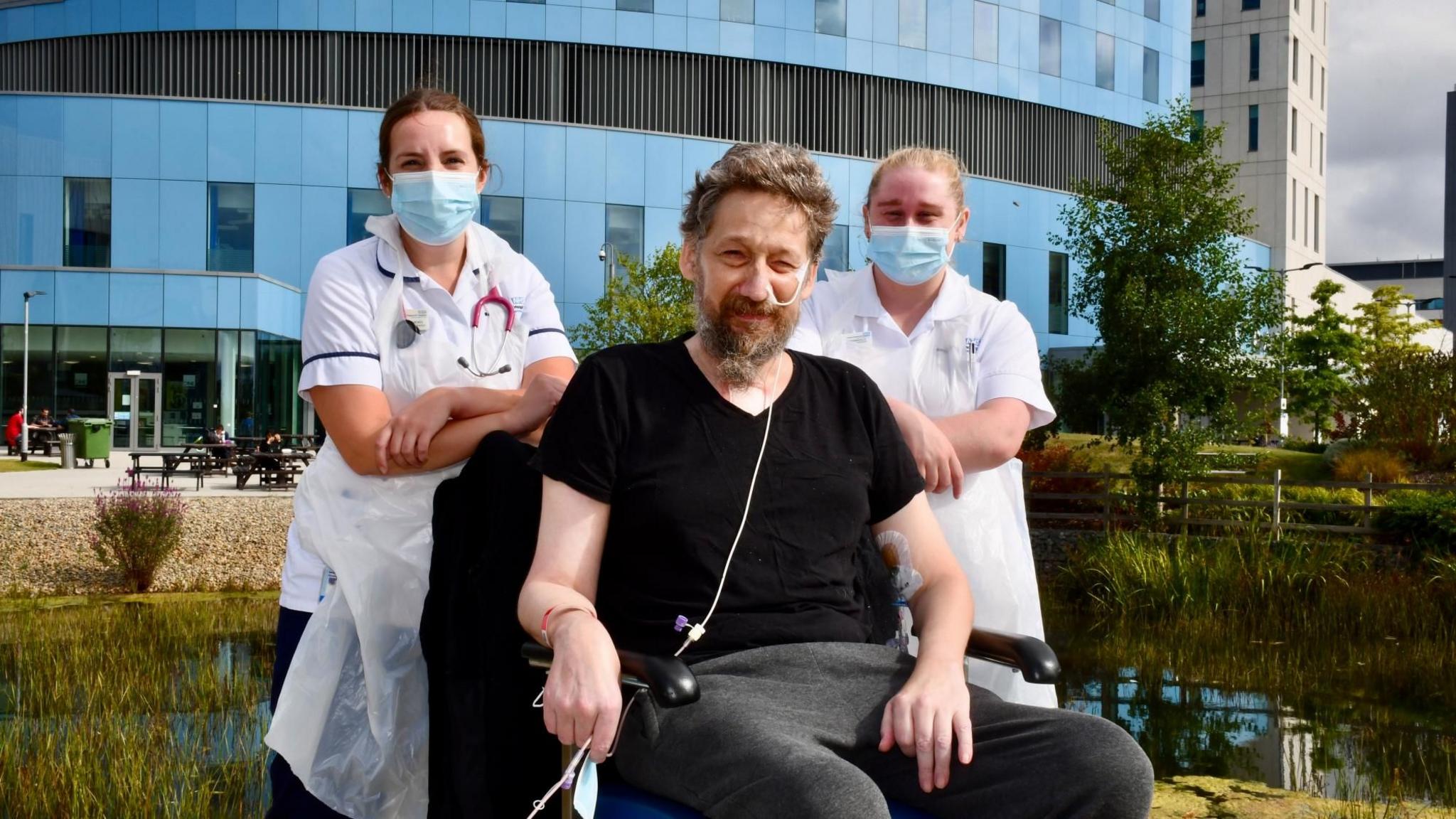 Daniel Evans-Smith sitting in a wheelchair outside Royal Papworth hospital. He is looking at the camera and has two female staff members standing behind him in white uniform. They are wearing face masks. Mr Evans-Smith has an oxygen tube going into his nose and taped to the side of his face. He is wearing a black t shirt and grey jogging bottoms.