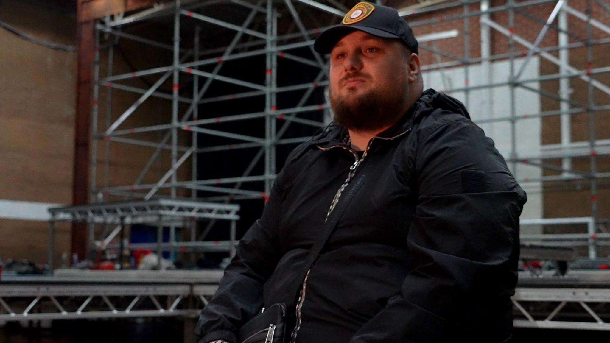 A man with a beard sitting down on the stage set with scaffolding in the background. He has a beard and is wearing a black cap with a badge on and a zip-up black jacket. 