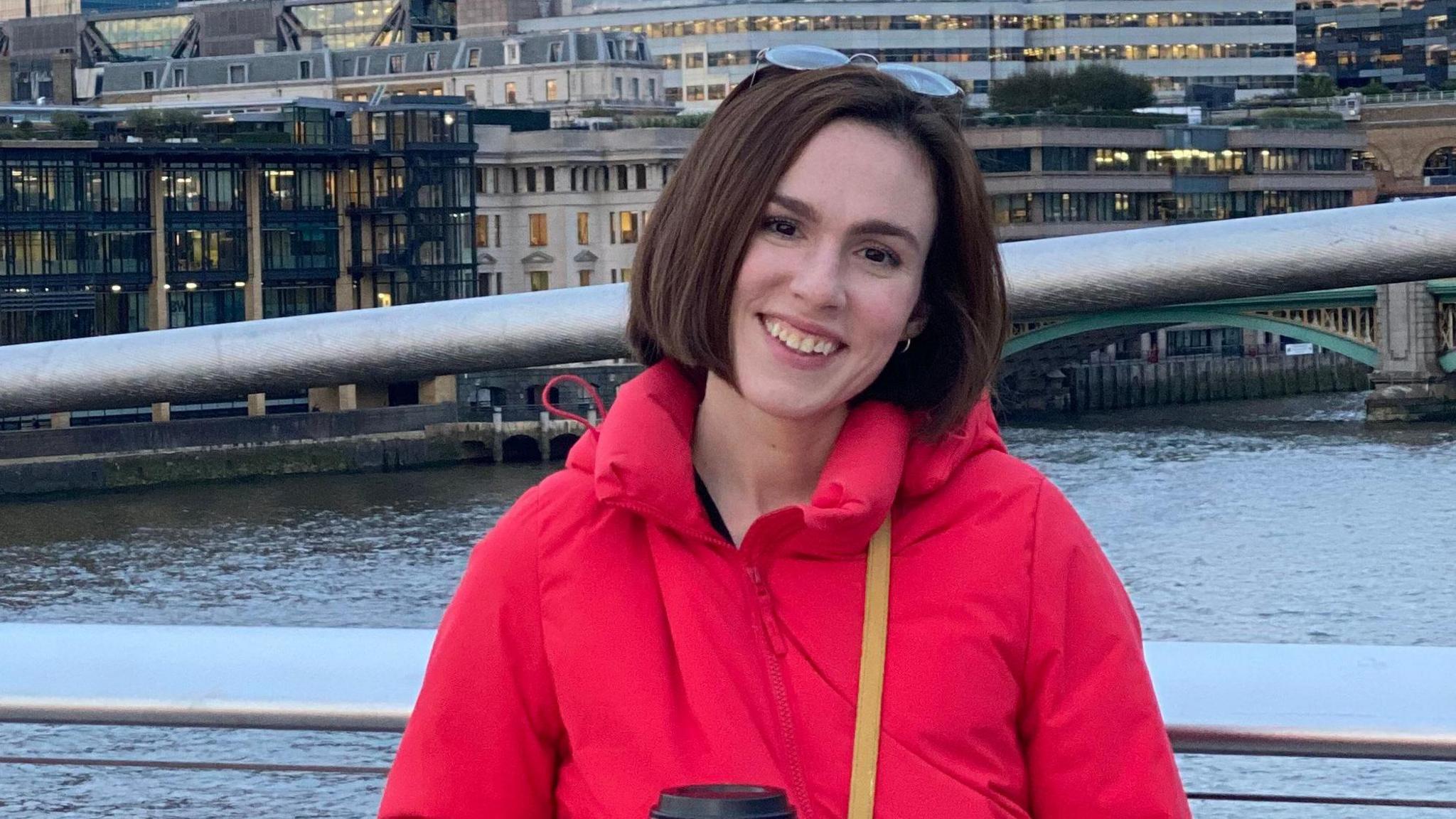 Emma Yates is smiling and wearing a red coat. She stands on a bridge over a river. 