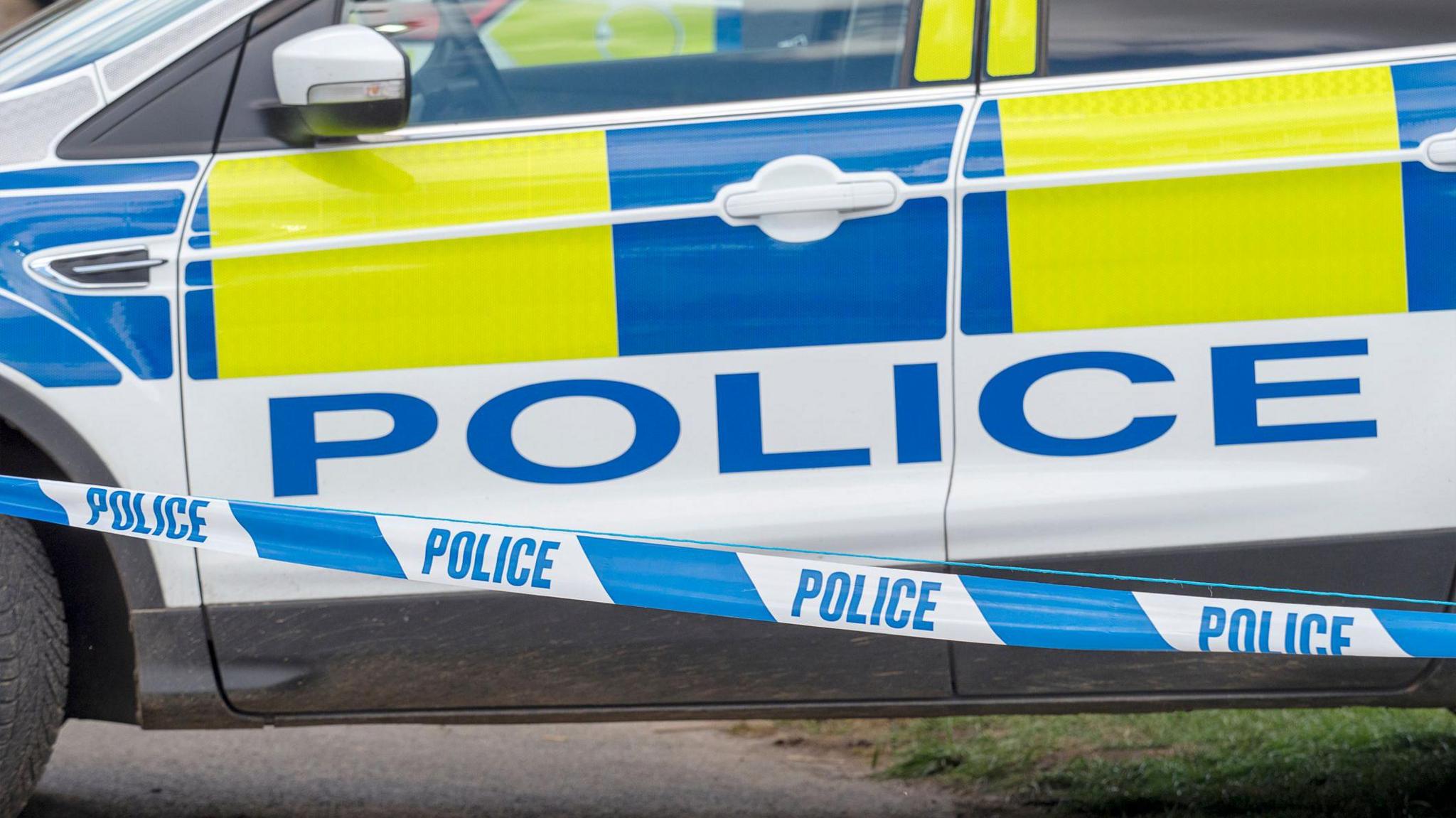 A police car with hi-vis yellow and blue decals parked on a grassy bank behind blue and white police cordon tape. 