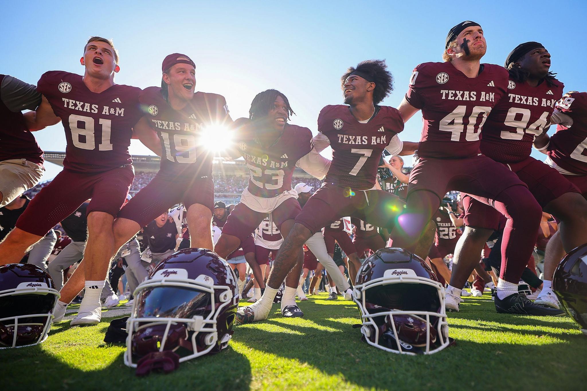American football players in maroon Texas A&M uniforms standing in a row with arms linked, helmets placed on the grass in front, sunlight shining through the stadium in the background.