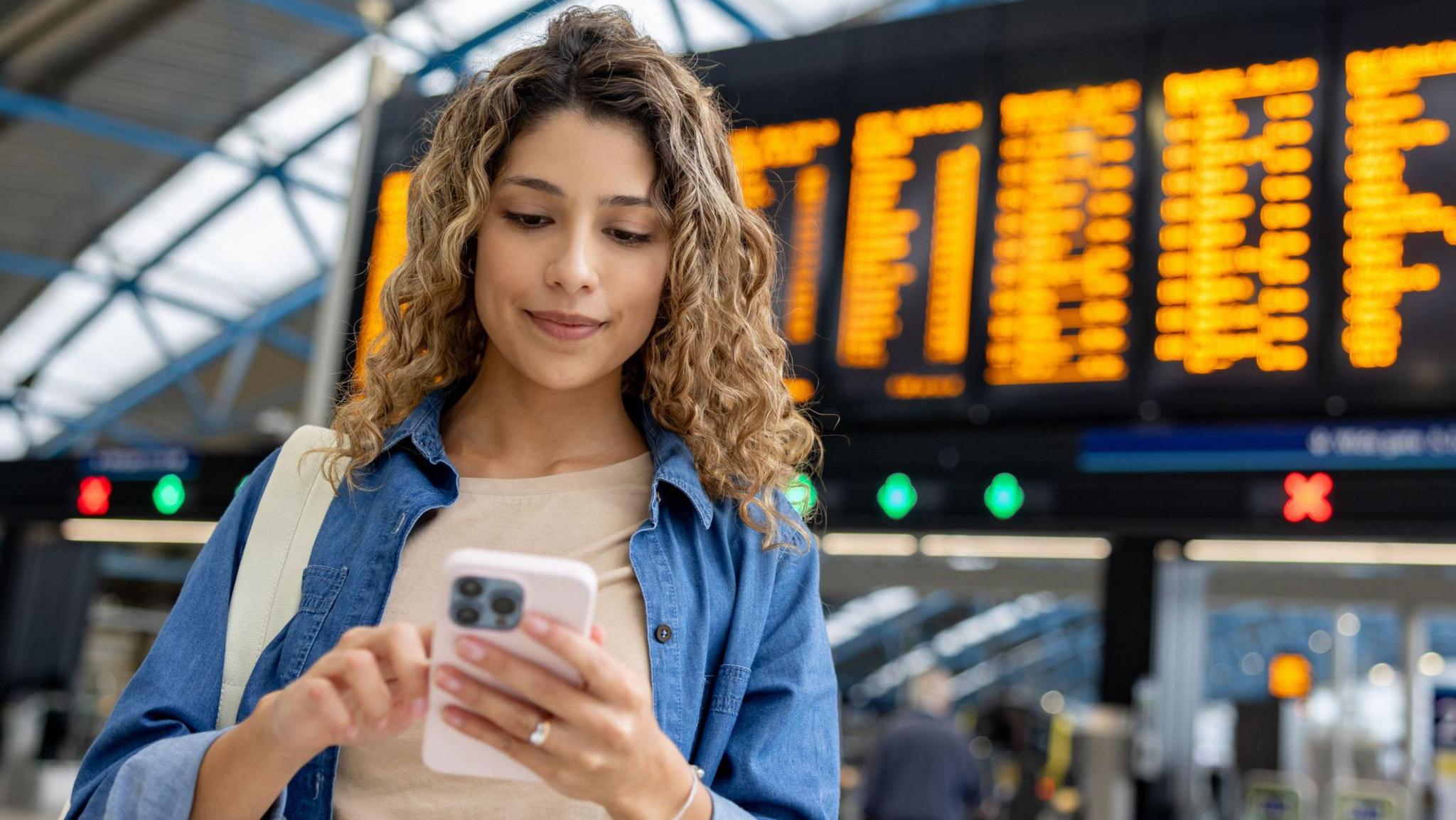 A woman buying ticket on phone at train station with arrival and departure boards in the background