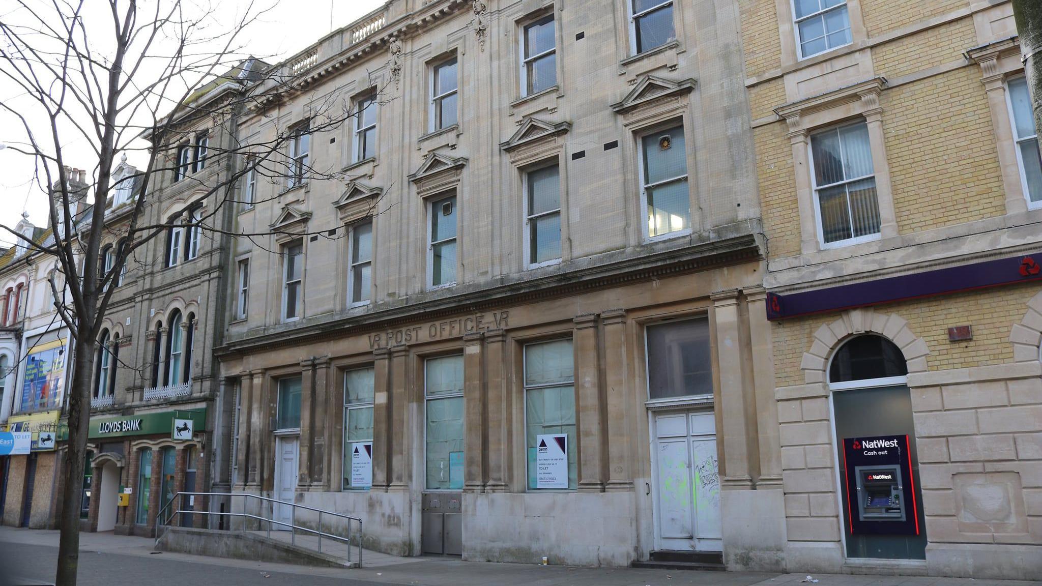 A view of the exterior of the former Lowestoft post office. The yellow brick building's lower windows and doors have been boarded up. The words post office can still be seen above the entrance.