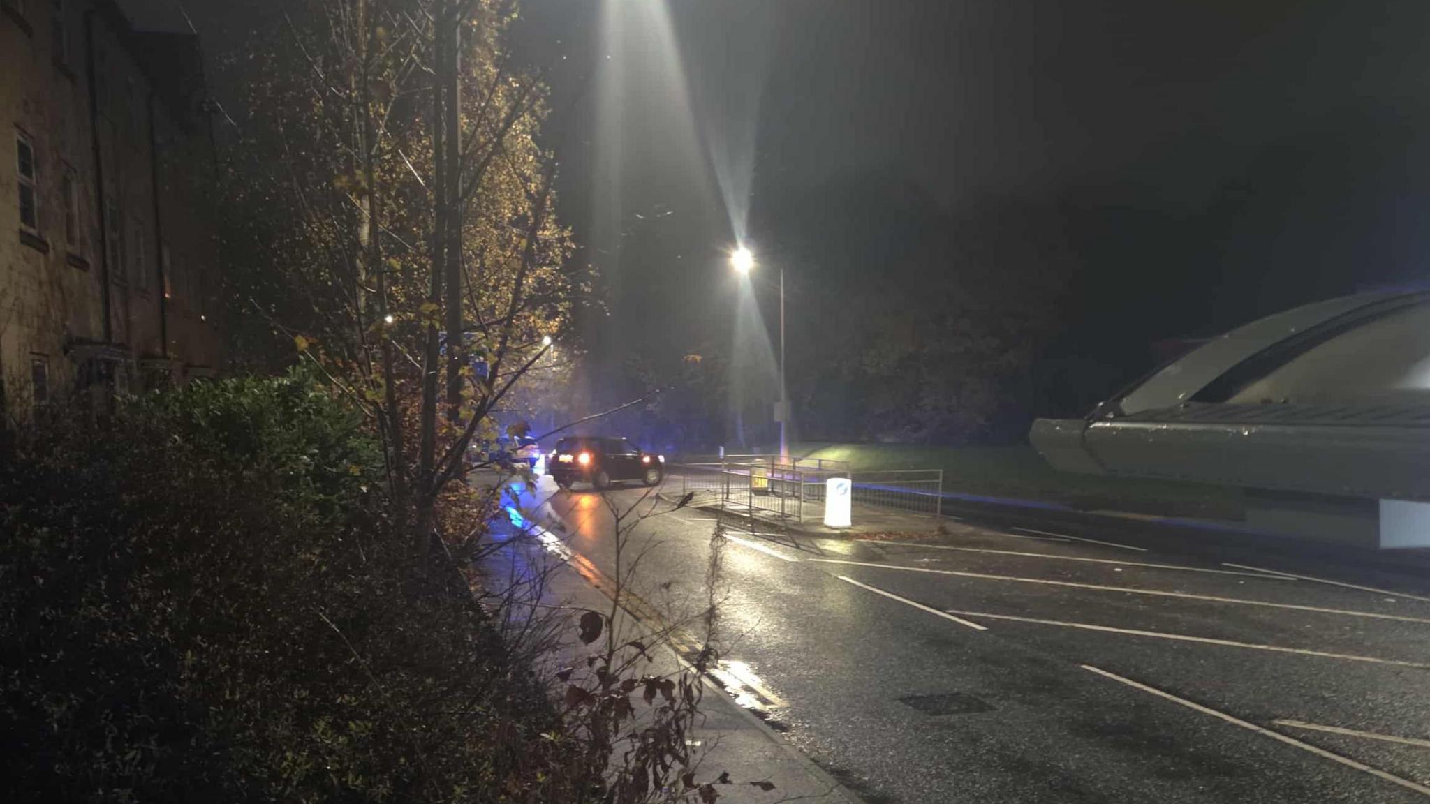 Road lit by streetlights, with a car in the background in the middle of the road with its lights on and a police car in the far distance.