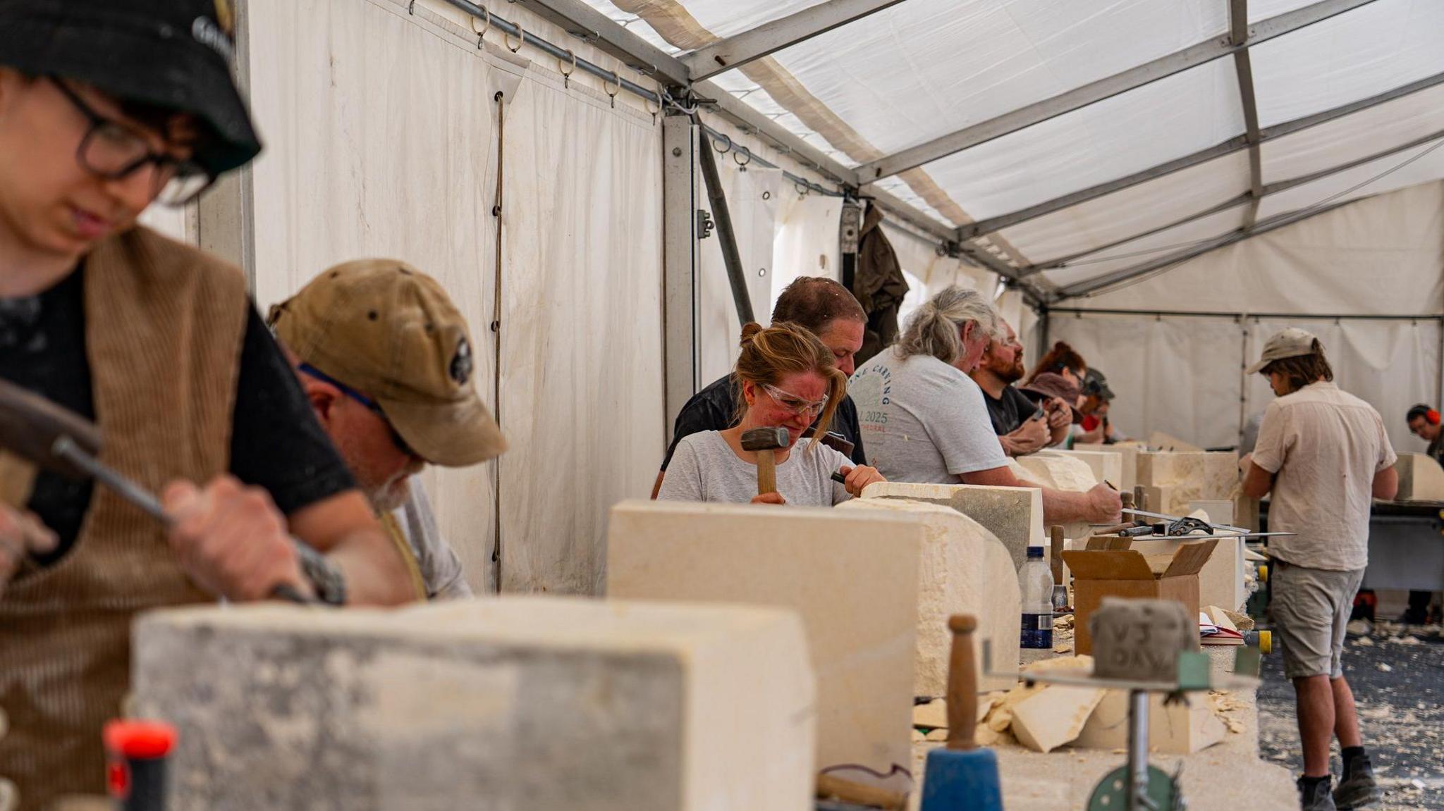 Looking down a row of stone carvers active working on blocks of stone with hammers and chisels. They all wear glasses or goggles and are concentrating. They are in a marquee.