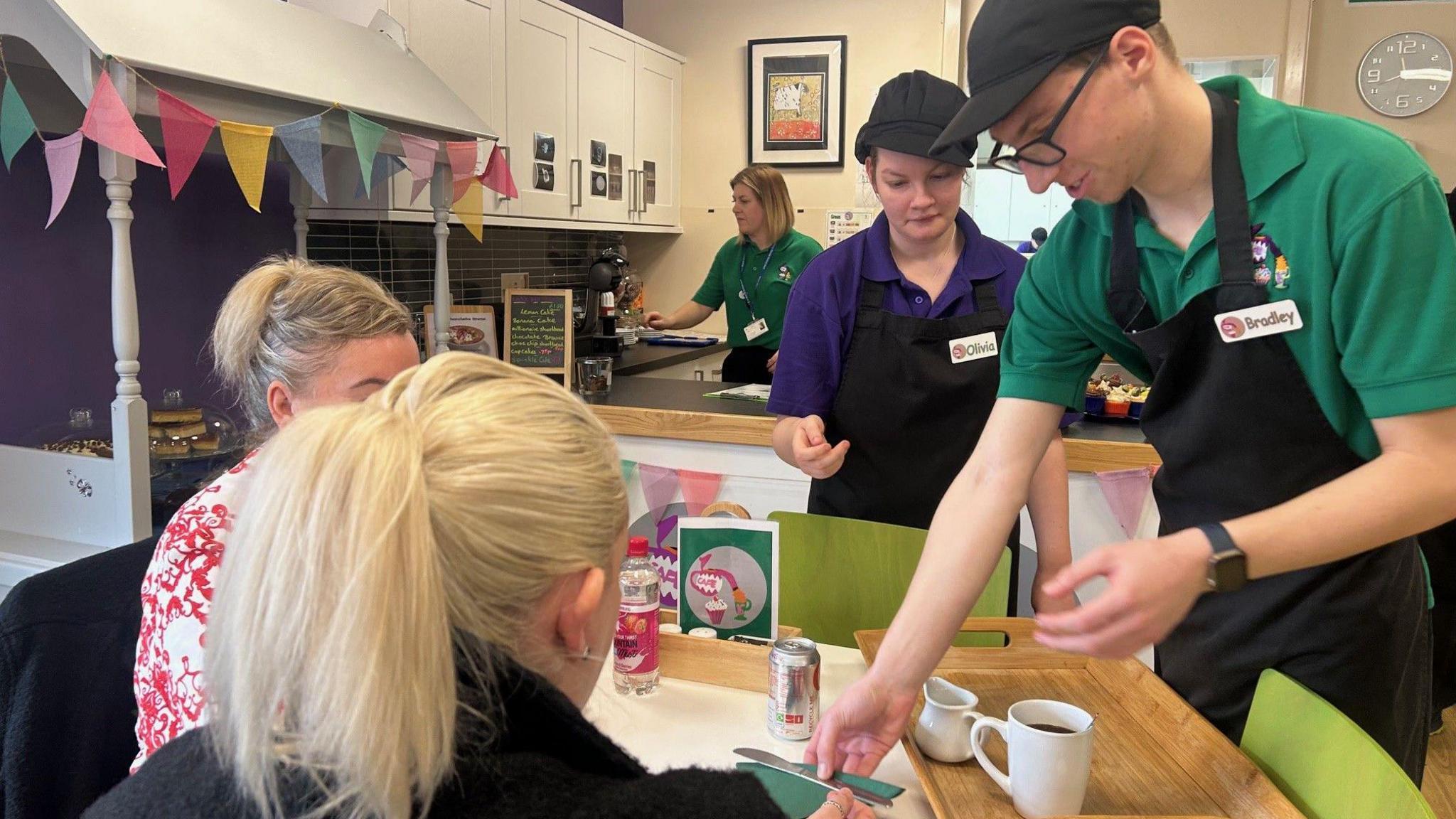 Two students from Priory Woods School serving customers in their community cafe. Bradley is one of the students and his name is displayed on his black apron.