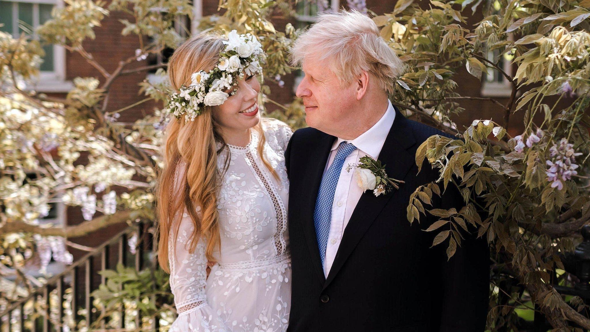 Boris Johnson standing next to his wife Carrie with his arm around her. He is wearing a black suit, white shirt and blue tie. Carrie is wearing a lace wedding dress with intricate detailing down the middle and on the sleeves. She is also wearing a white rose flower crown and smiling at her husband. They are standing on an outdoor staircase next to a bush with pink flowers on it. 