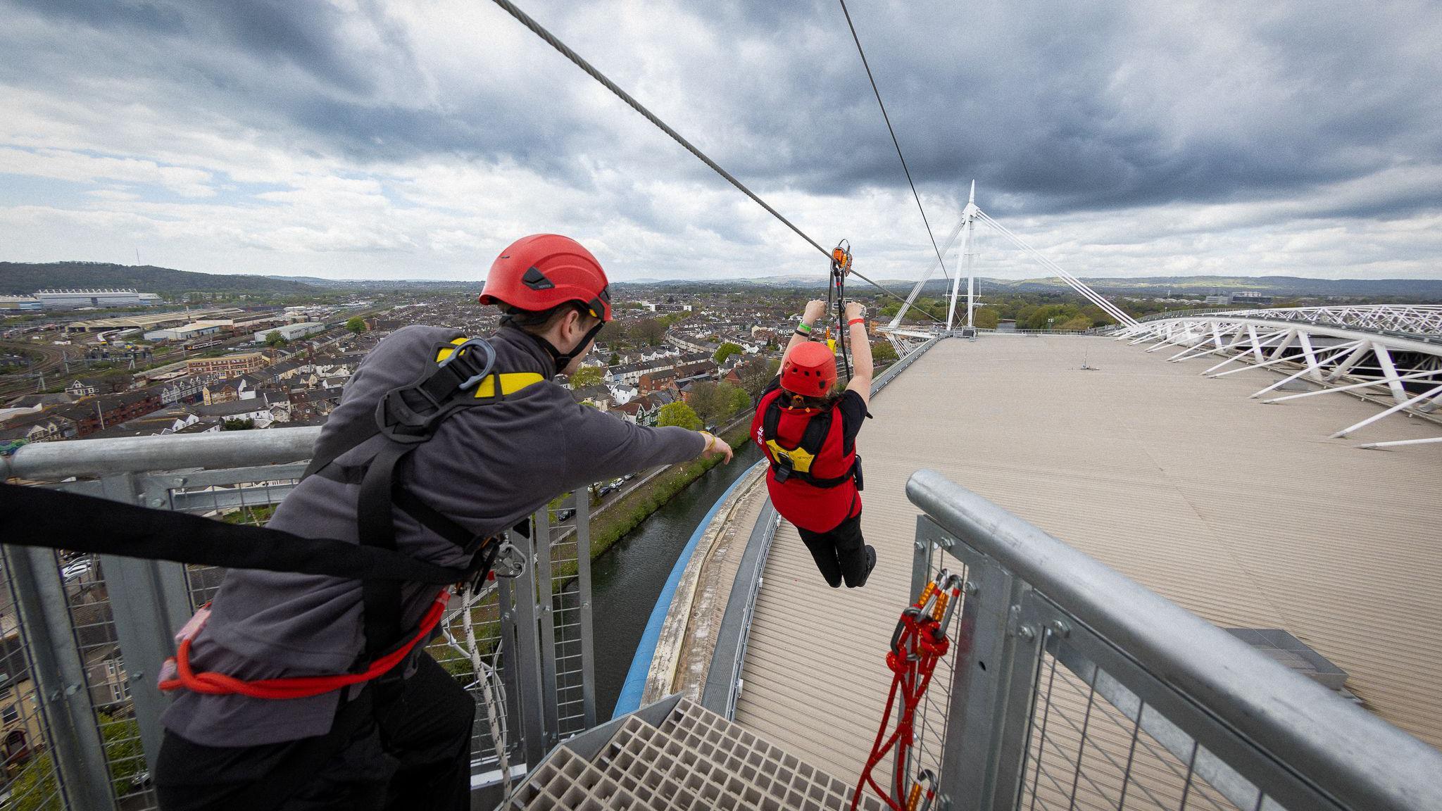 The top of the principality stadium from a platform used to launch the zip liner. A man on the left of the photo is strapped on with a black strap. His arm is outstretched towards a woman who has just launched. He is hanging on to a handle next to the zip line and roller. She is strapped in with a shoulder harness visible. The roof of the stadium and the city beyond are in the background.