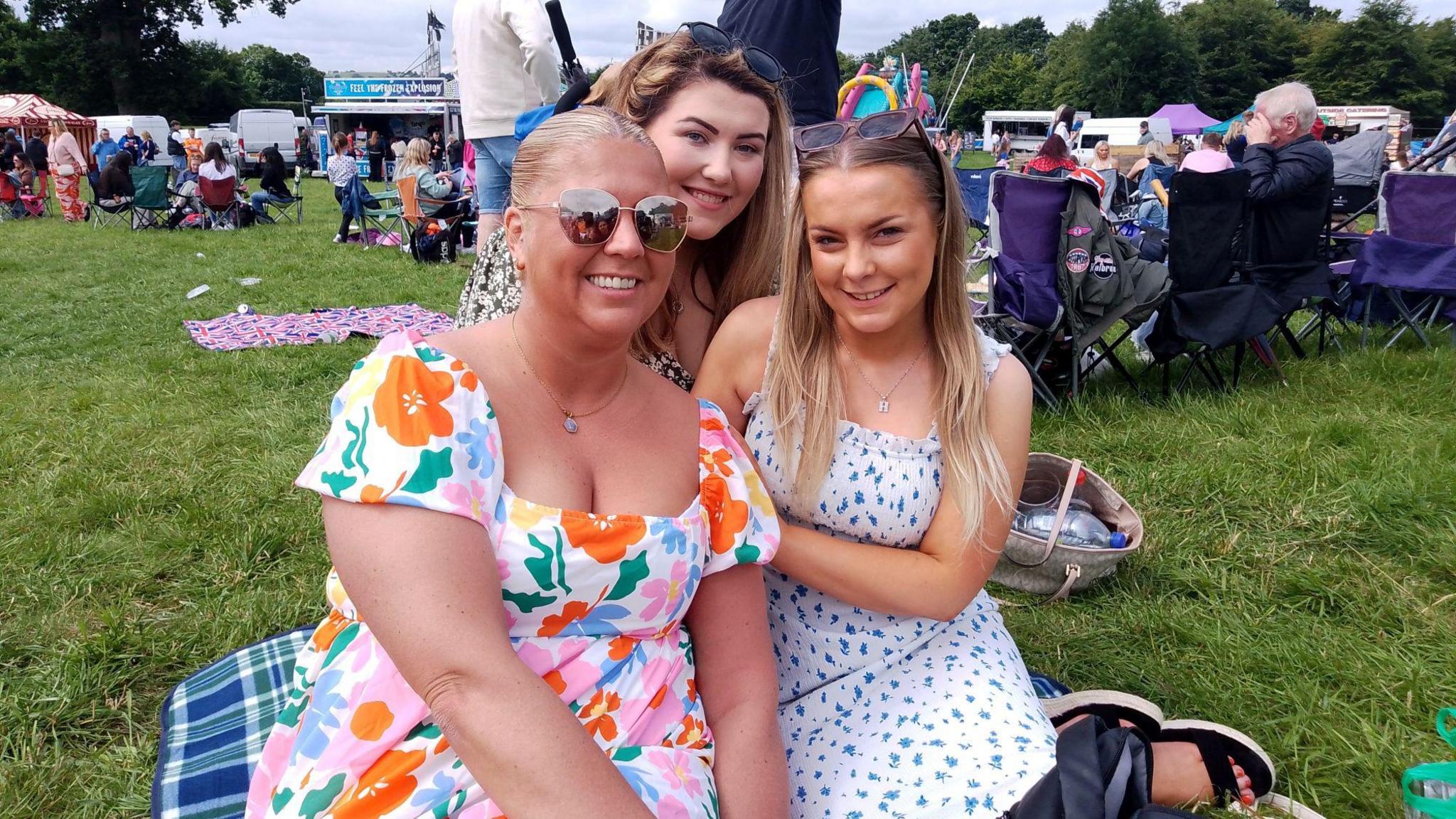 A blonde woman wearing a multi-colour floral patterned dress sits on a picnic blanket, behind her is a dark haired woman smiling and also sitting on the blanket is blonde woman wearing a white and blue floral dress and sunglasses.