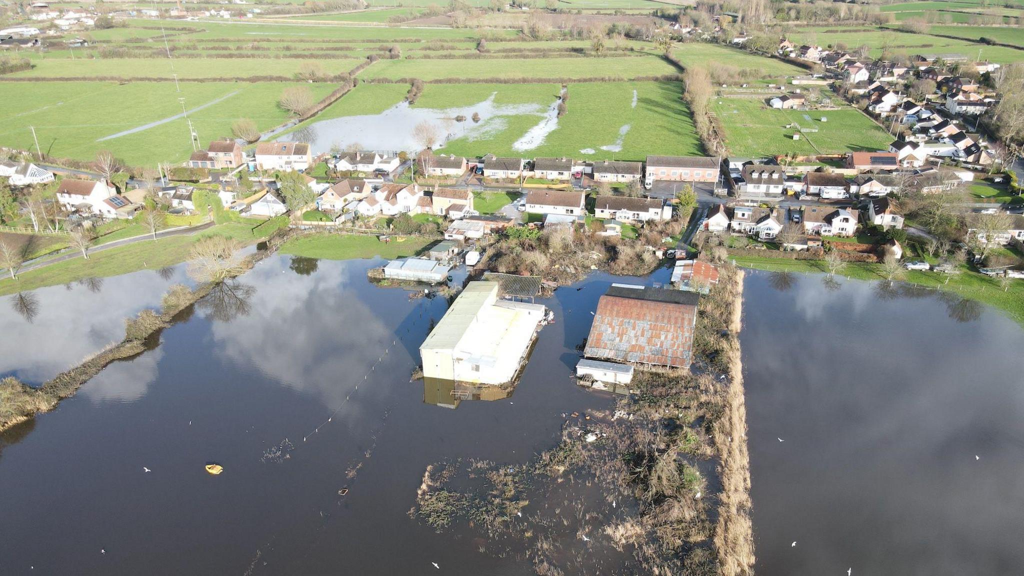 A drone shot of houses surrounded by fields. The fields are flooded and water is surrounding a number of buildings.
