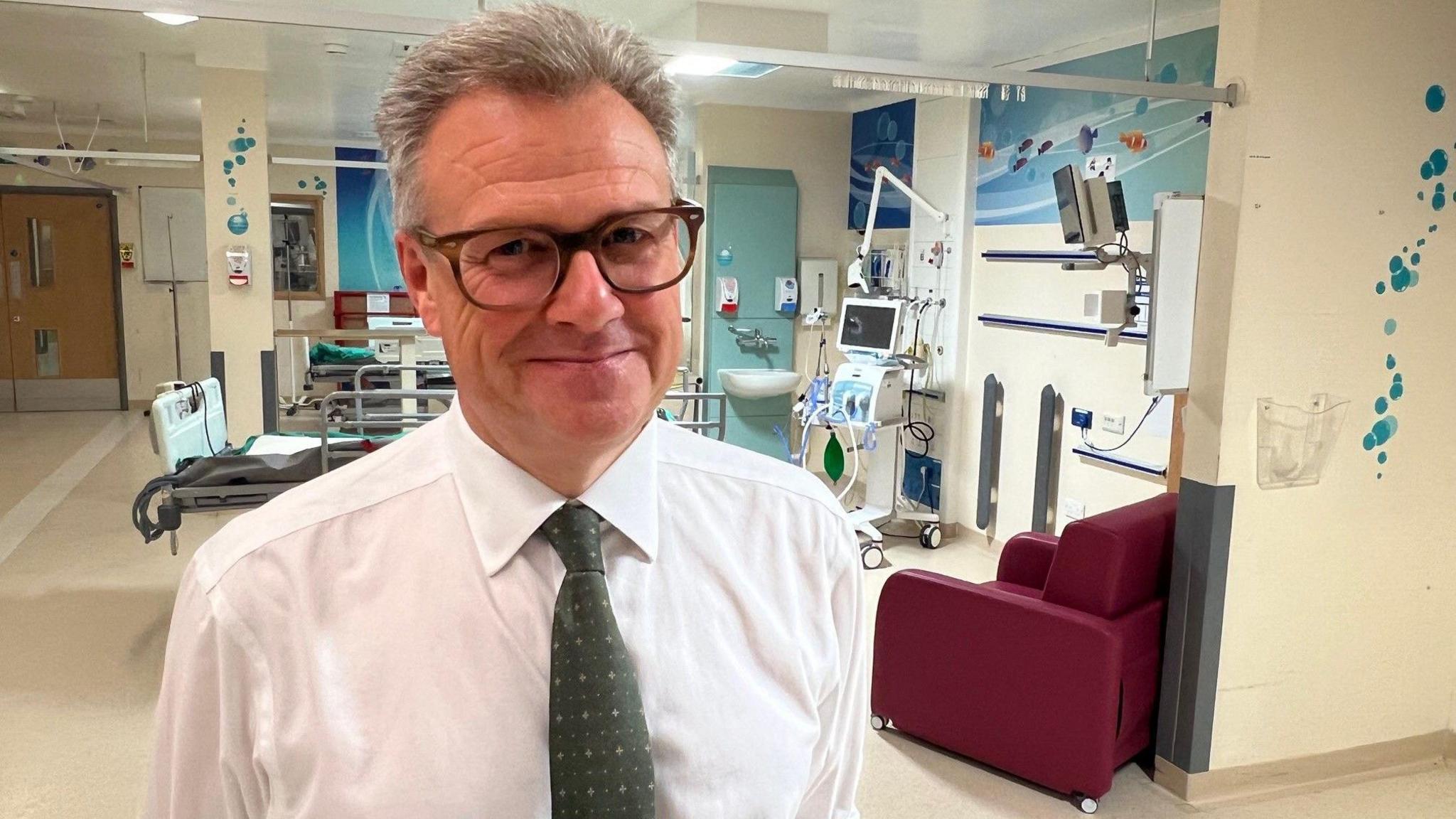 A white man with grey hair and dark glasses, wearing a white shirt and tie stands in front of hospital equipment 